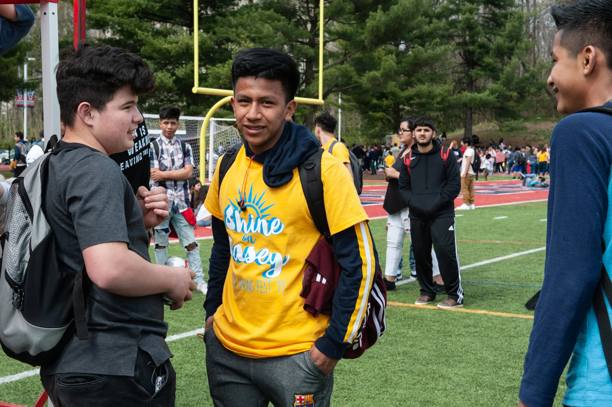 Young boys standing and talking on a school athletic field, with more students in the background, some walking and some gathered in groups.