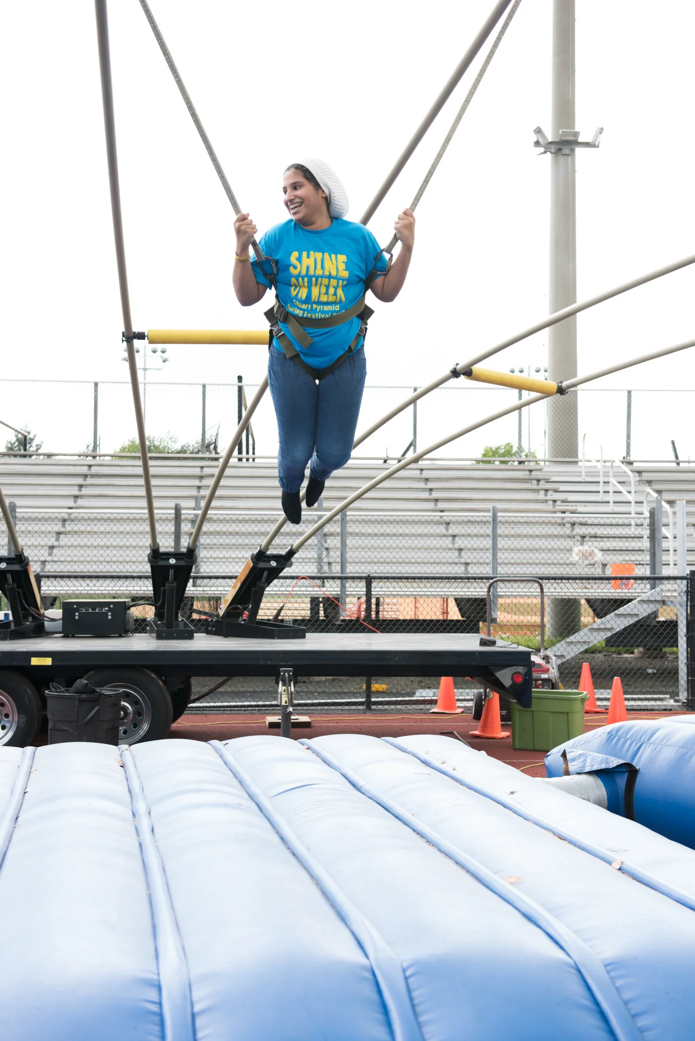 A woman wearing a blue UK Shine On Week t-shirt and a white headband bouncing on a trampoline at an outdoor event, with bleachers and a fence in the background.