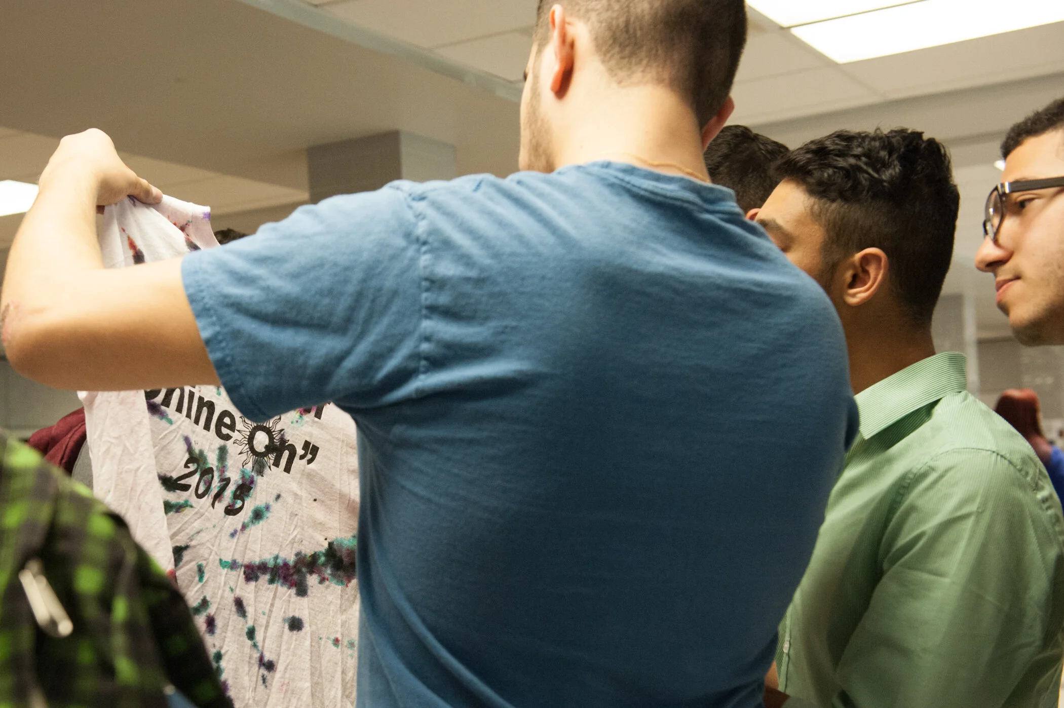 Group of young men in a room, one holding a painted shirt with text, others observing.