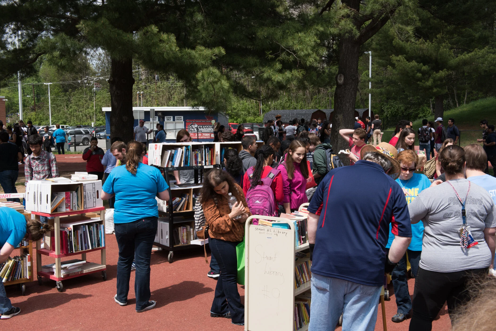 People browsing books at an outdoor book sale on a school track field, with trees and parked cars in the background.