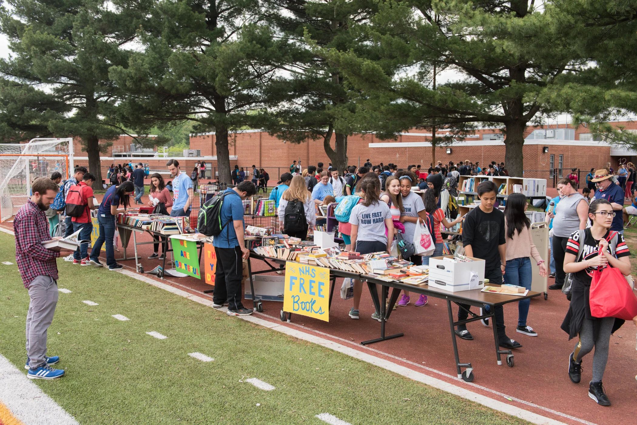 Students walking past tables of free books at an outdoor book sale on a school track, with trees and a school building in the background.