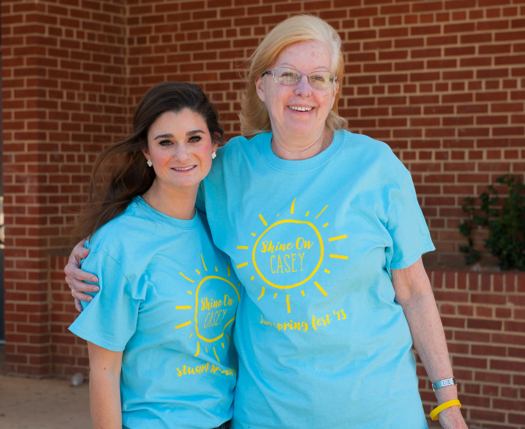 Two women smiling and standing outdoors in front of a brick wall, wearing matching turquoise t-shirts with yellow text and graphics for a school event.