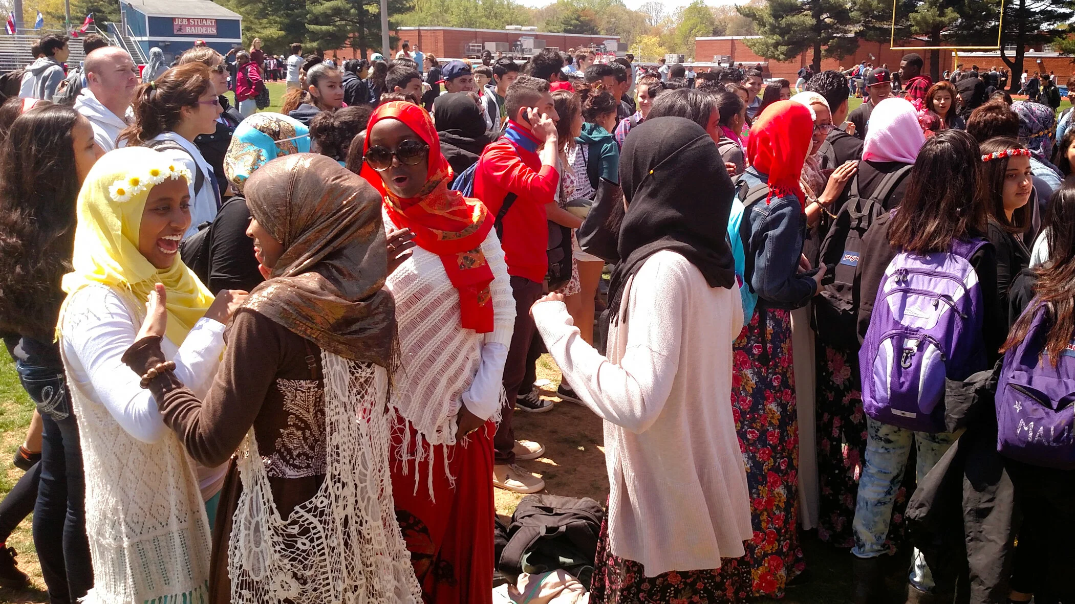 A large diverse group of students gathered outdoors on a school field, some wearing headscarves, engaging in conversations with a background of school buildings and a football field.