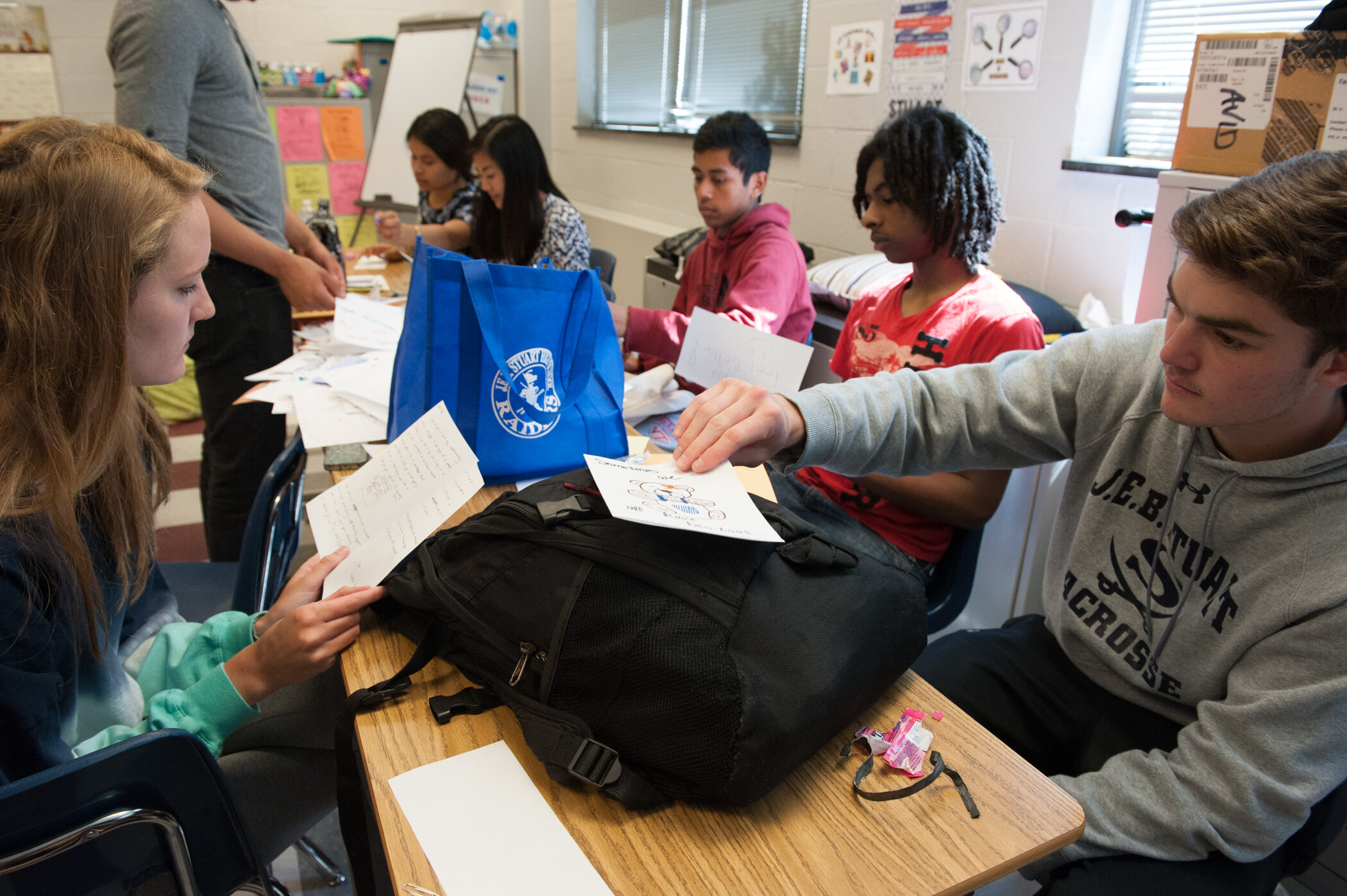 Students sitting at a classroom table exchanging papers, with backpacks and supplies visible, in a classroom setting.