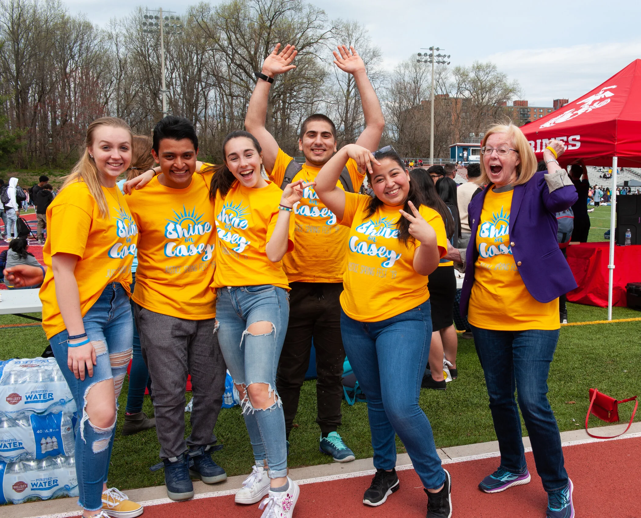 Group of young people smiling and having fun at an outdoor event, wearing matching yellow T-shirts with blue writing, on a track field with trees, a red tent, and water bottles in the background.