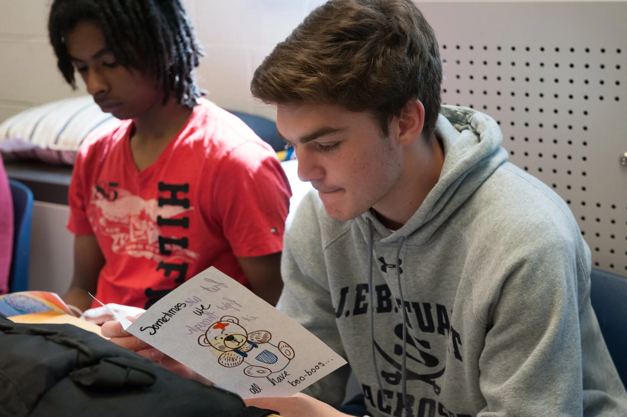 Two teenage boys sitting at a table, one reading a handmade card with a teddy bear illustration and the message "Sometimes you just have to be yourself. You have all the boo-boos".