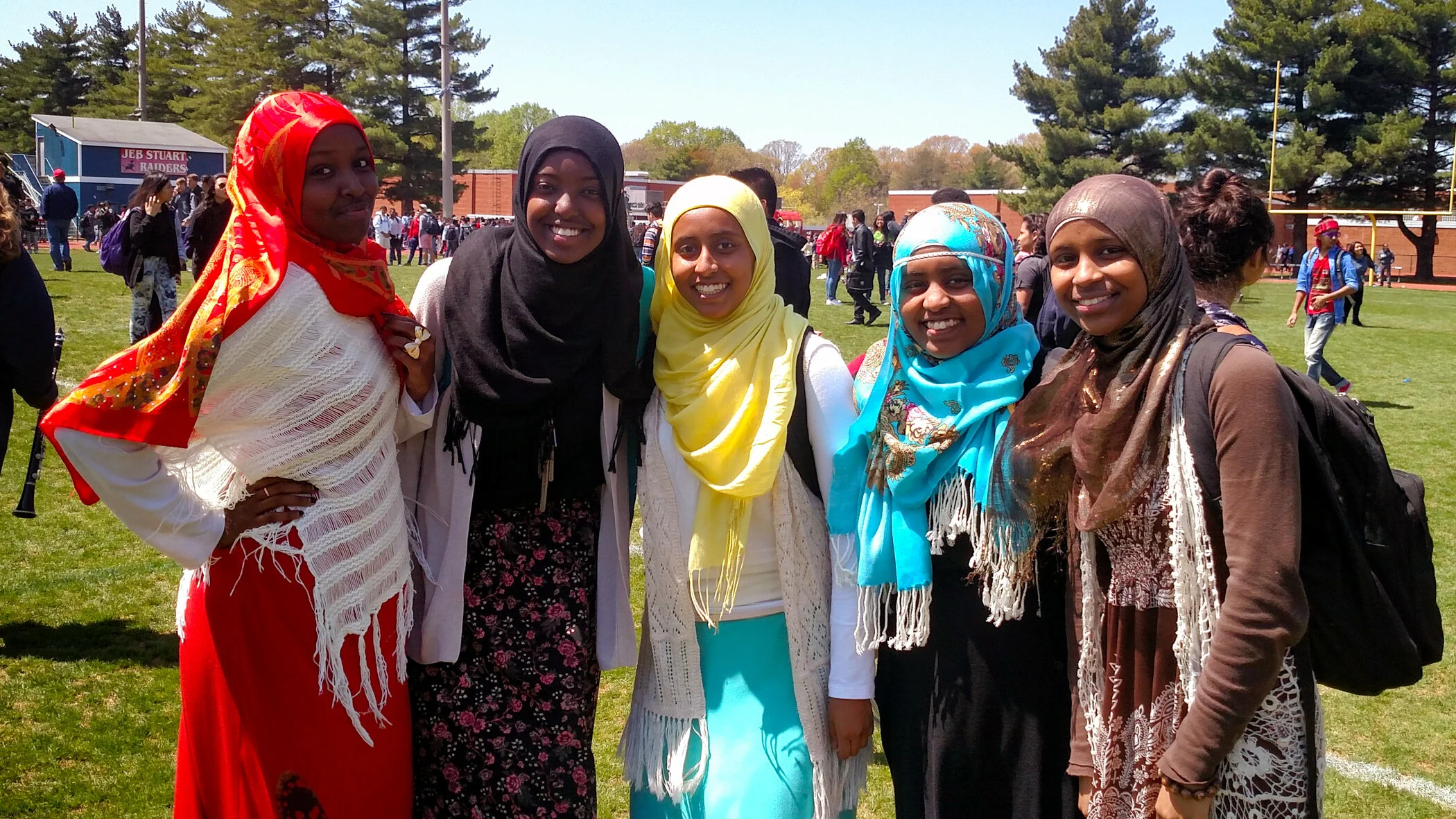 Five women wearing hijabs, standing together on a school football field during daytime, smiling at the camera.