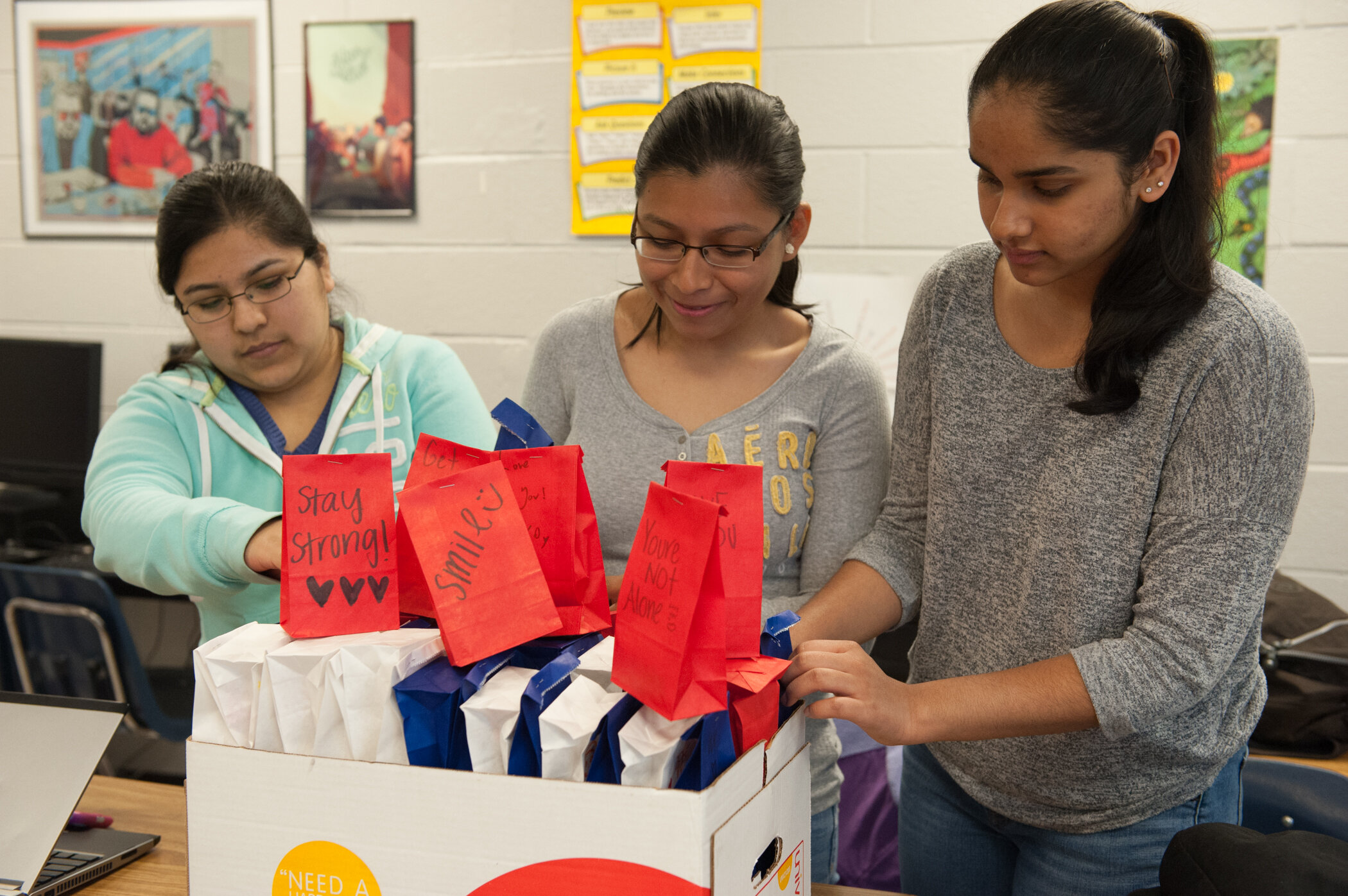 Three young women are standing around a cardboard box filled with paper packages, some with handwritten messages like 'Stay Strong!', 'Smile! :)', and 'You're Not Alone.' they are engaged in an activity together in a classroom decorated with colorful