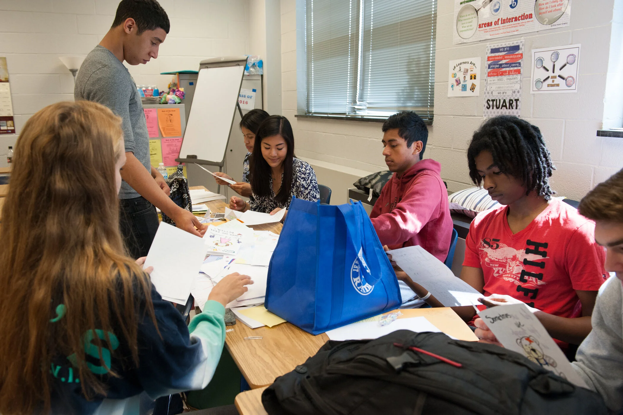 A teacher standing at a table full of students working on papers and drawings in a classroom.