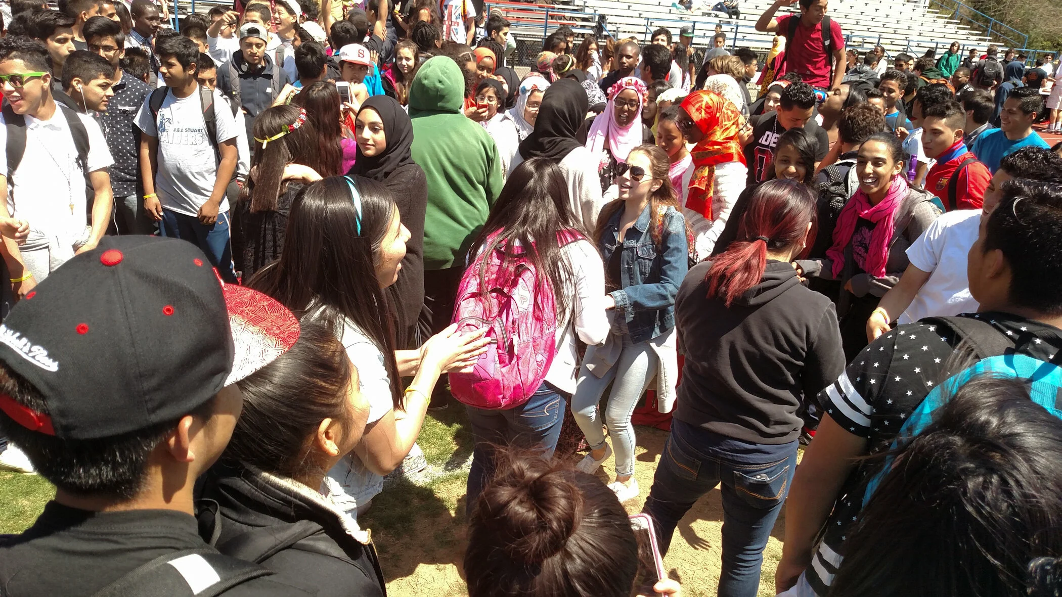 Crowd of students gathered outdoors, some talking and smiling, some wearing backpacks and sunglasses, on a sunny day at a school event or assembly.