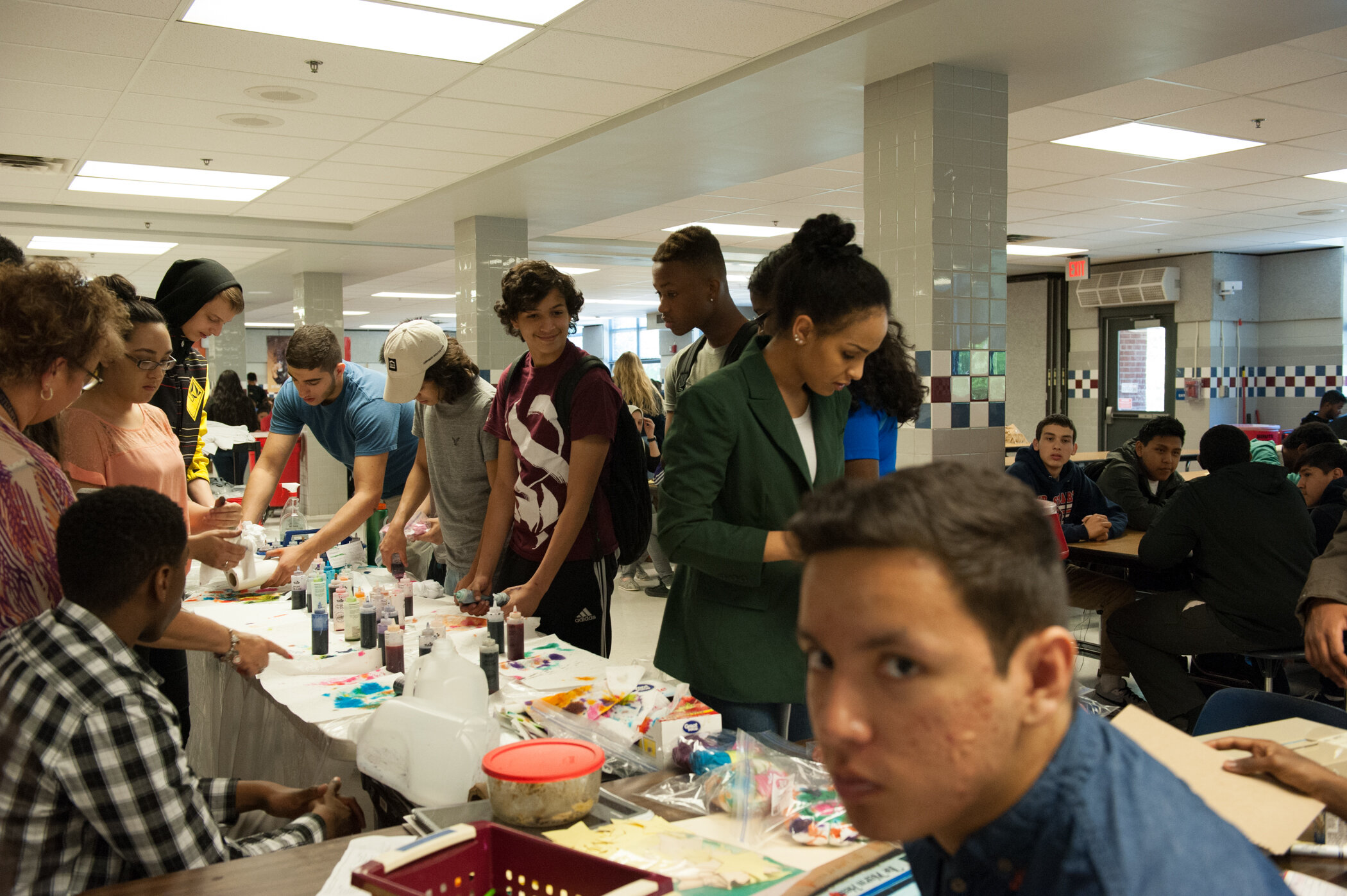 A group of young people at a table in a cafeteria or community center, engaging in arts and crafts activities with various supplies, while others sit at tables in the background.