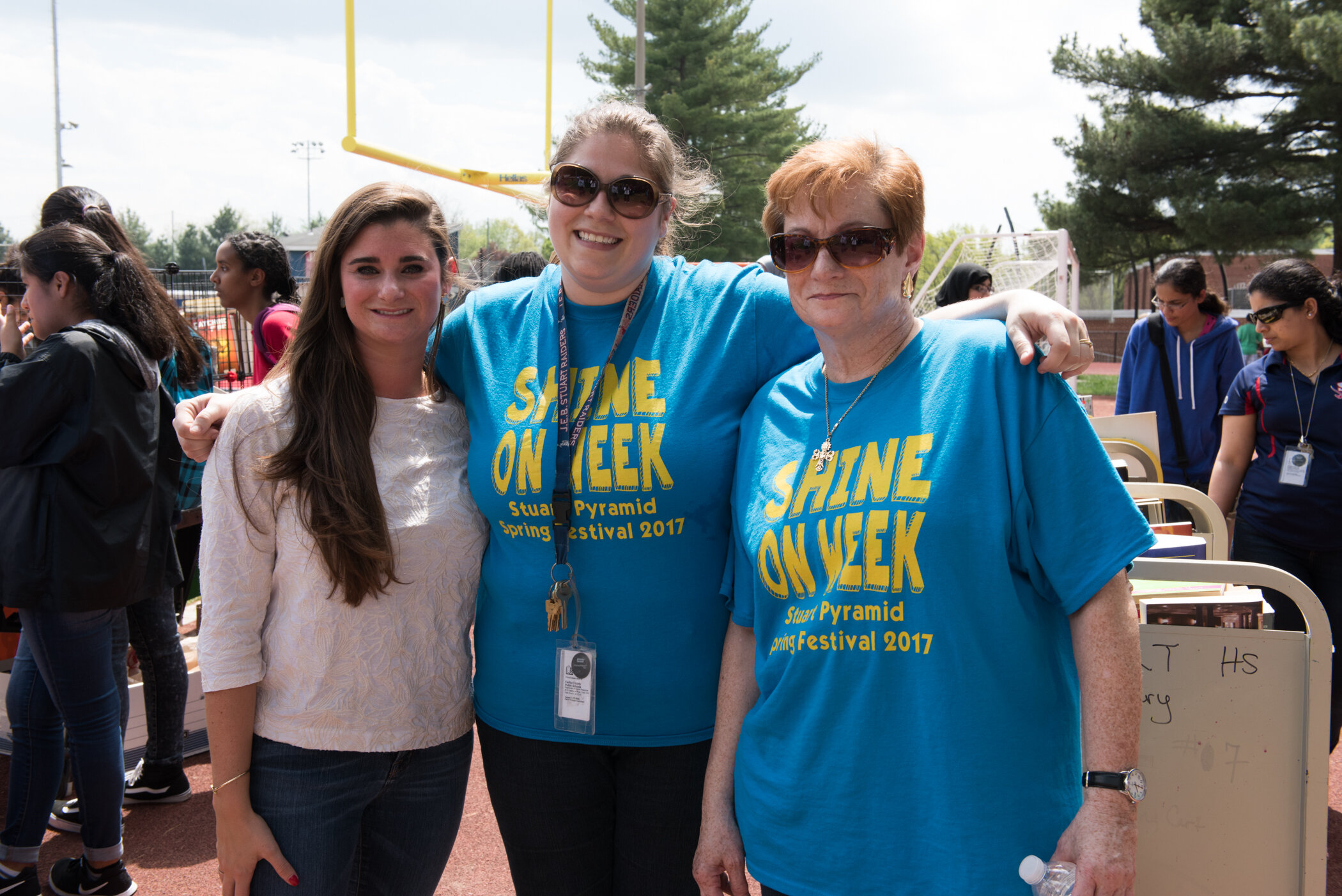Three women smiling and posing together outdoors at a school event. Two are wearing matching blue T-shirts with yellow text that says "SHINE ON WEEK" and "Spring Festival 2017." The woman on the left is wearing a white top and dark pants. The backgro