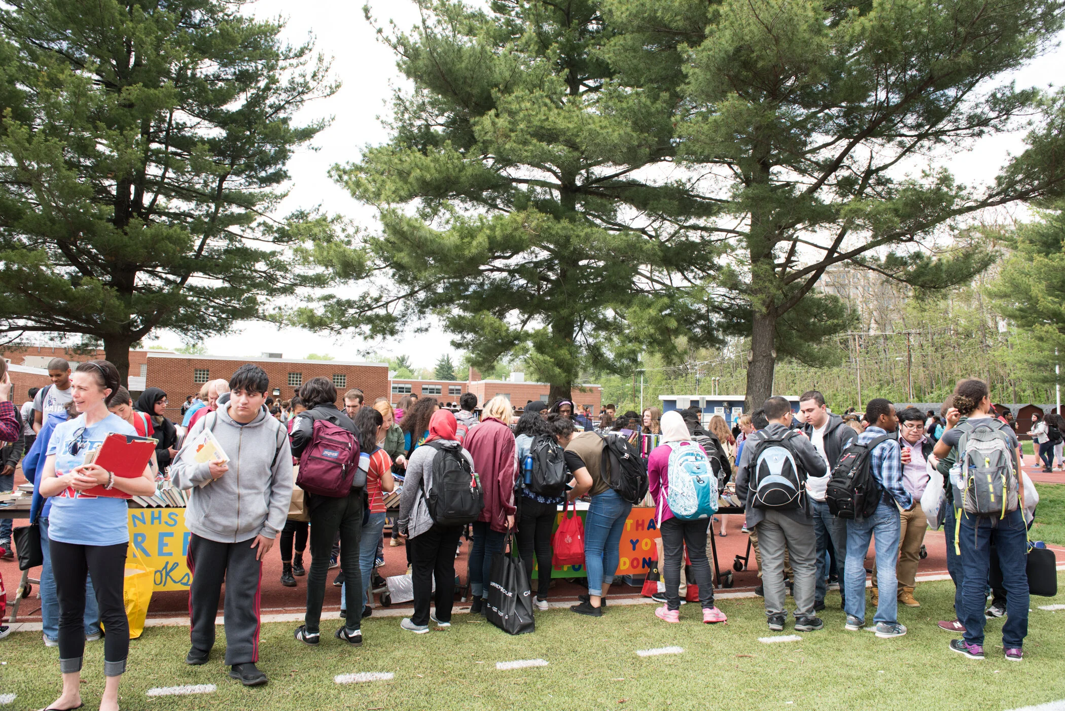 A large group of students gathered outdoors, standing by tables with signs, on a grassy area with trees and a school building in the background.