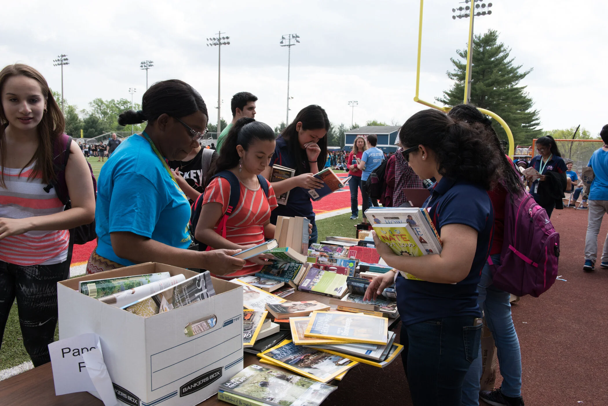 People browsing books at an outdoor book sale on a sports field with a football goalpost in the background, overcast sky, and some trees.