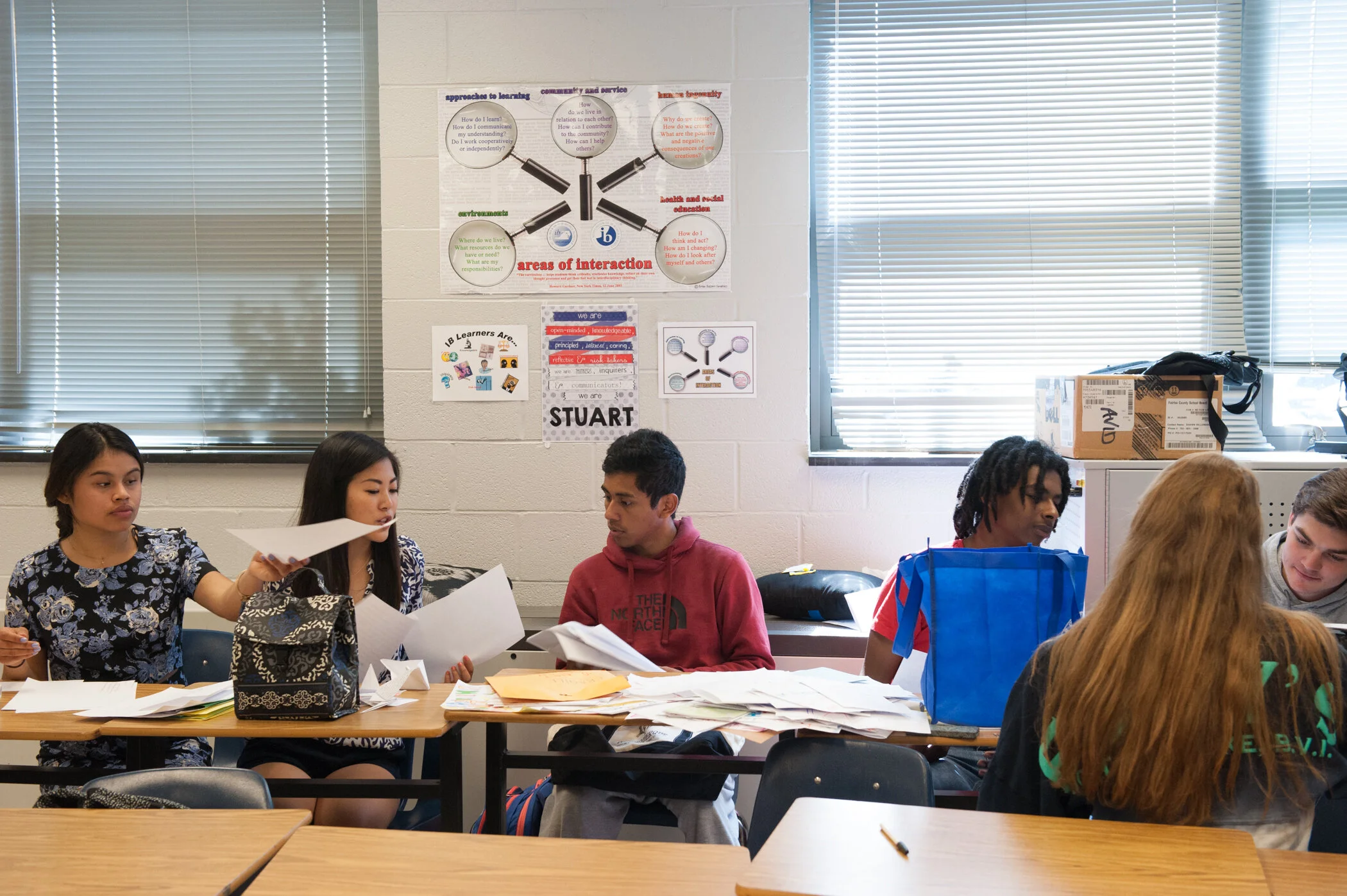 Students sitting at desks in a classroom, working with papers and books, with posters on the wall behind them.