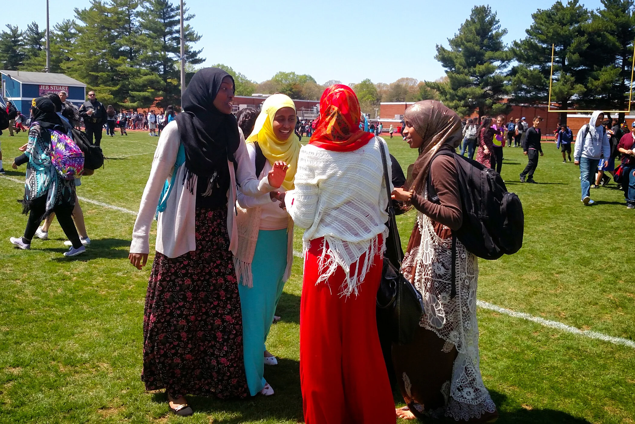 Group of women wearing colorful hijabs and dresses standing together on a grassy field during a sunny day, with students walking and socializing in the background.