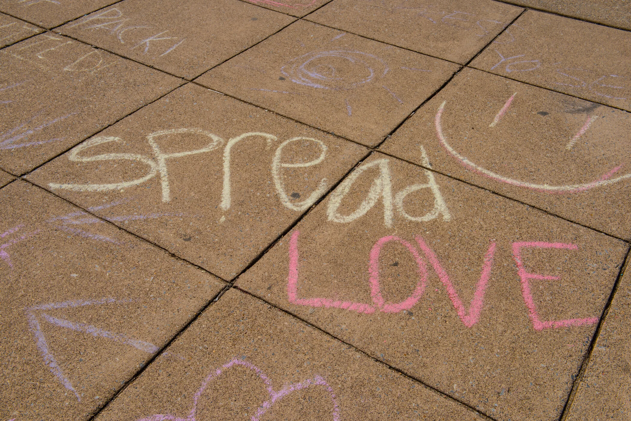 Colorful sidewalk chalk drawings on concrete sidewalk, including the words 'Spread Love' with a smiley face and hearts.