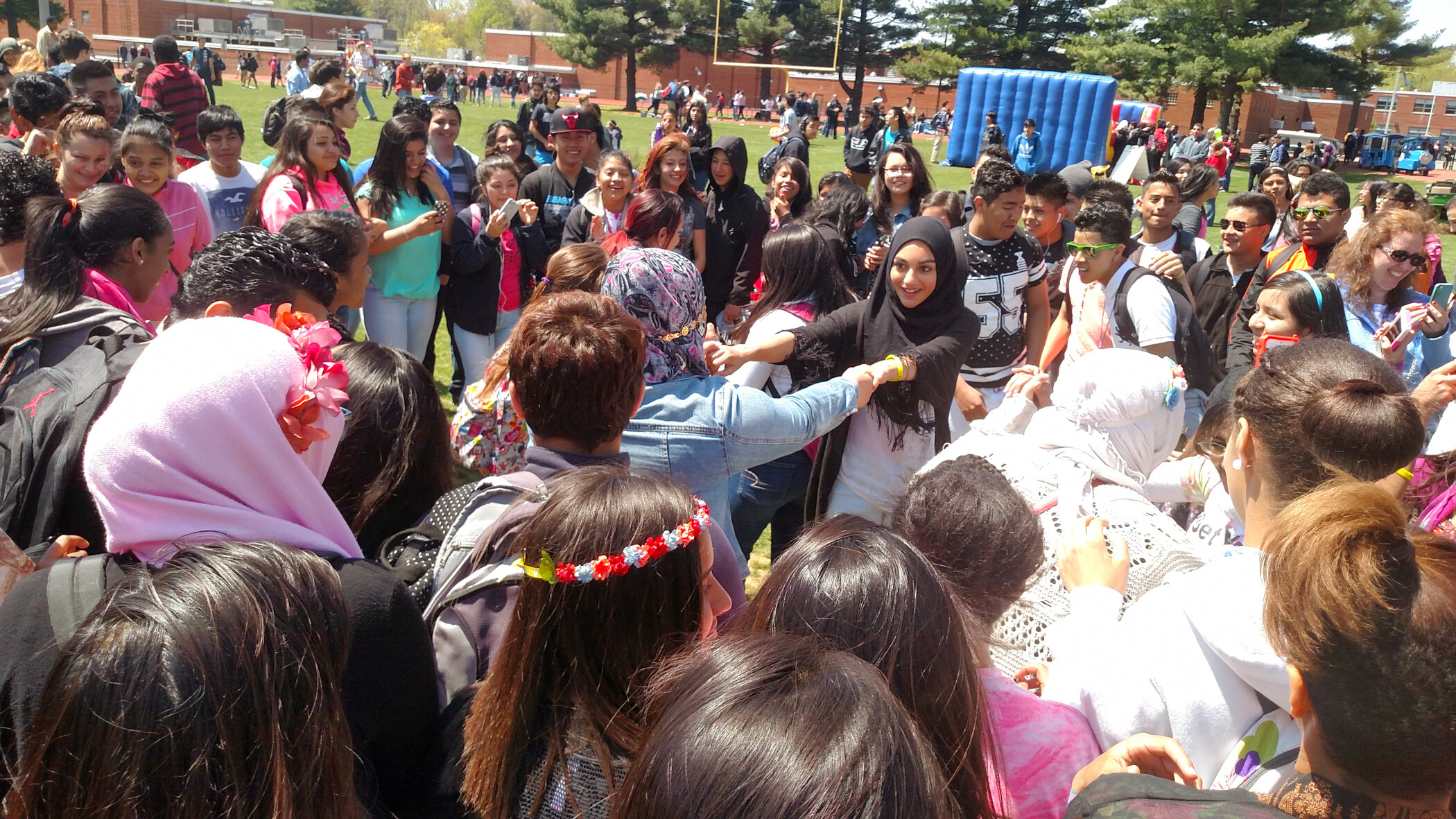 A large group of diverse students are outdoors on a school field, participating in a social activity, with some taking pictures and engaging with each other.