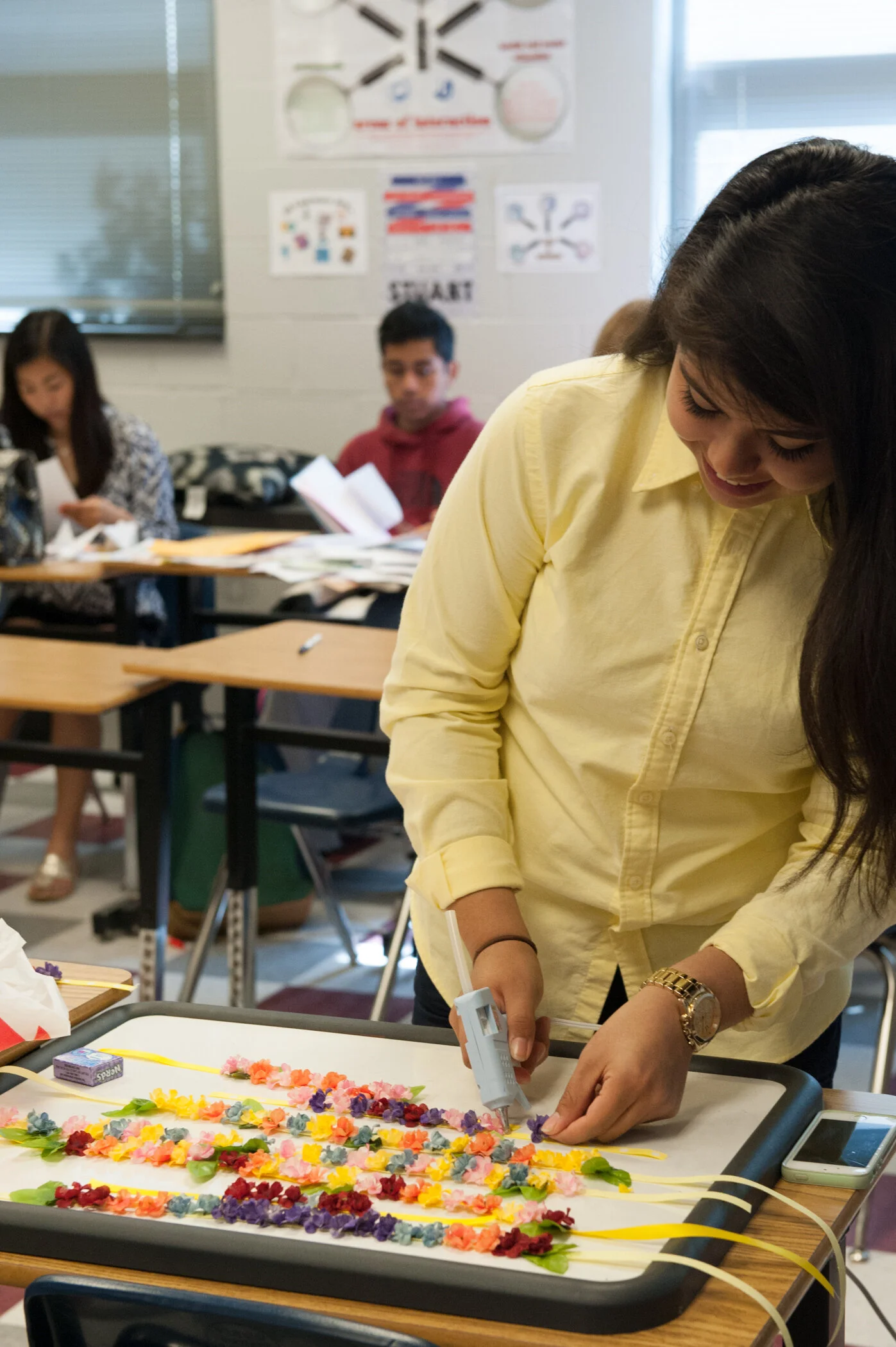 A girl in a yellow shirt working on a craft project with colorful paper flowers and a hot glue gun in a classroom.