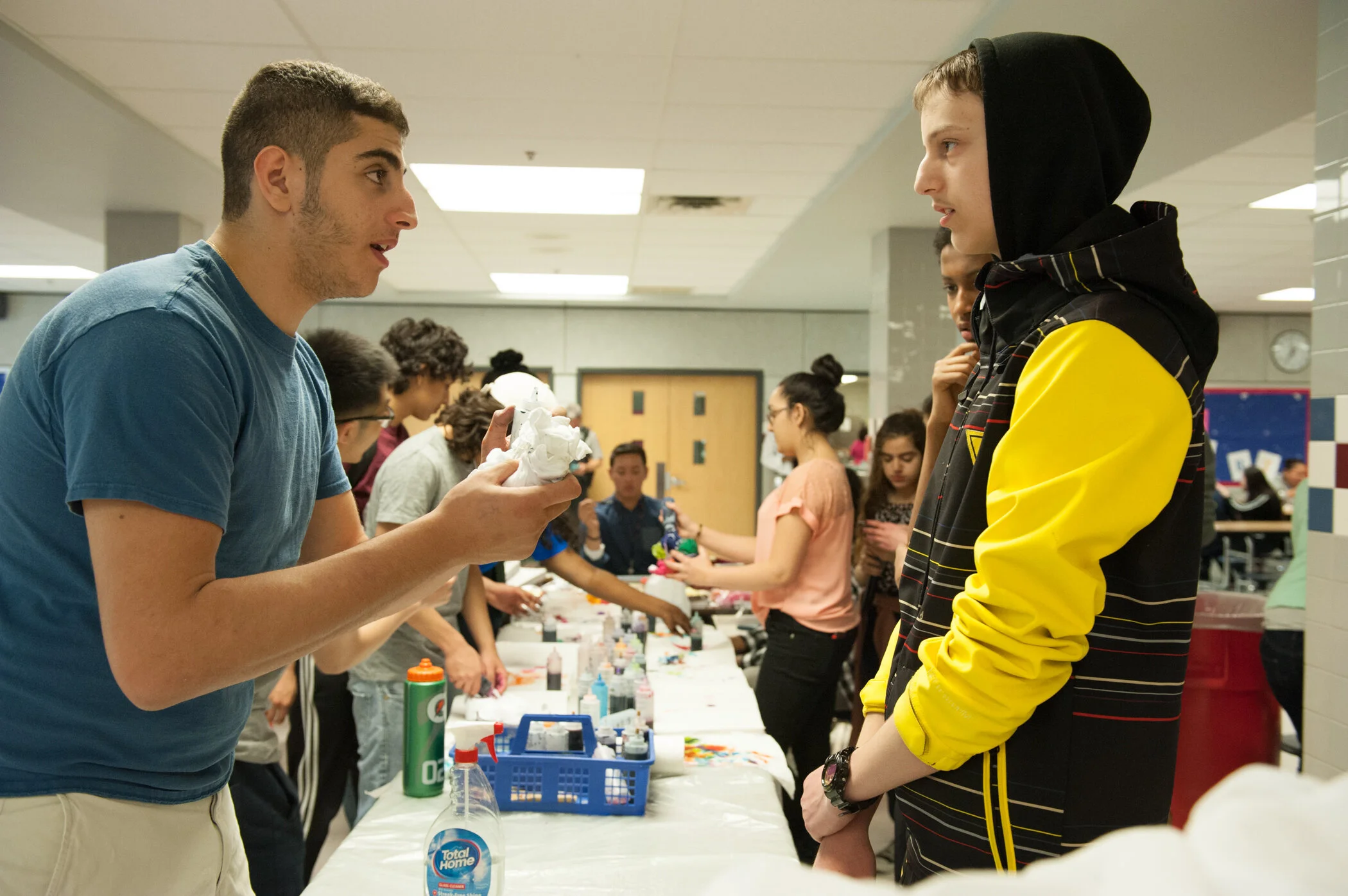 A young man in a blue t-shirt shows a crumpled piece of paper to another young man wearing a black hoodie with yellow sleeves, during a school event with students working on arts and crafts at a long table.