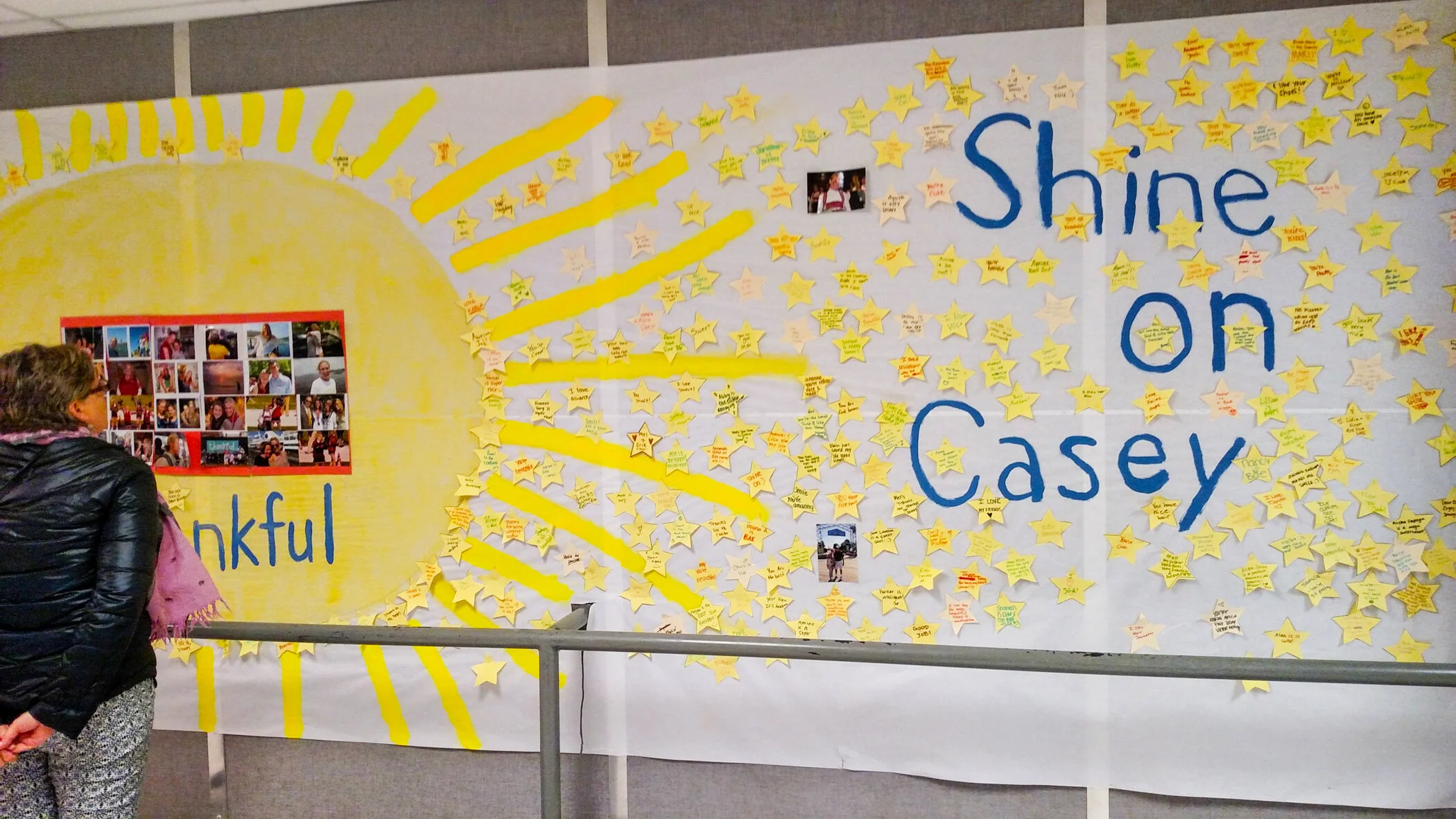 A woman observing a large display board with yellow star-shaped notes and photographs. The background features a yellow sun drawing with rays and the words "Shine On Casey" written in blue. The notes contain handwritten messages of appreciation.