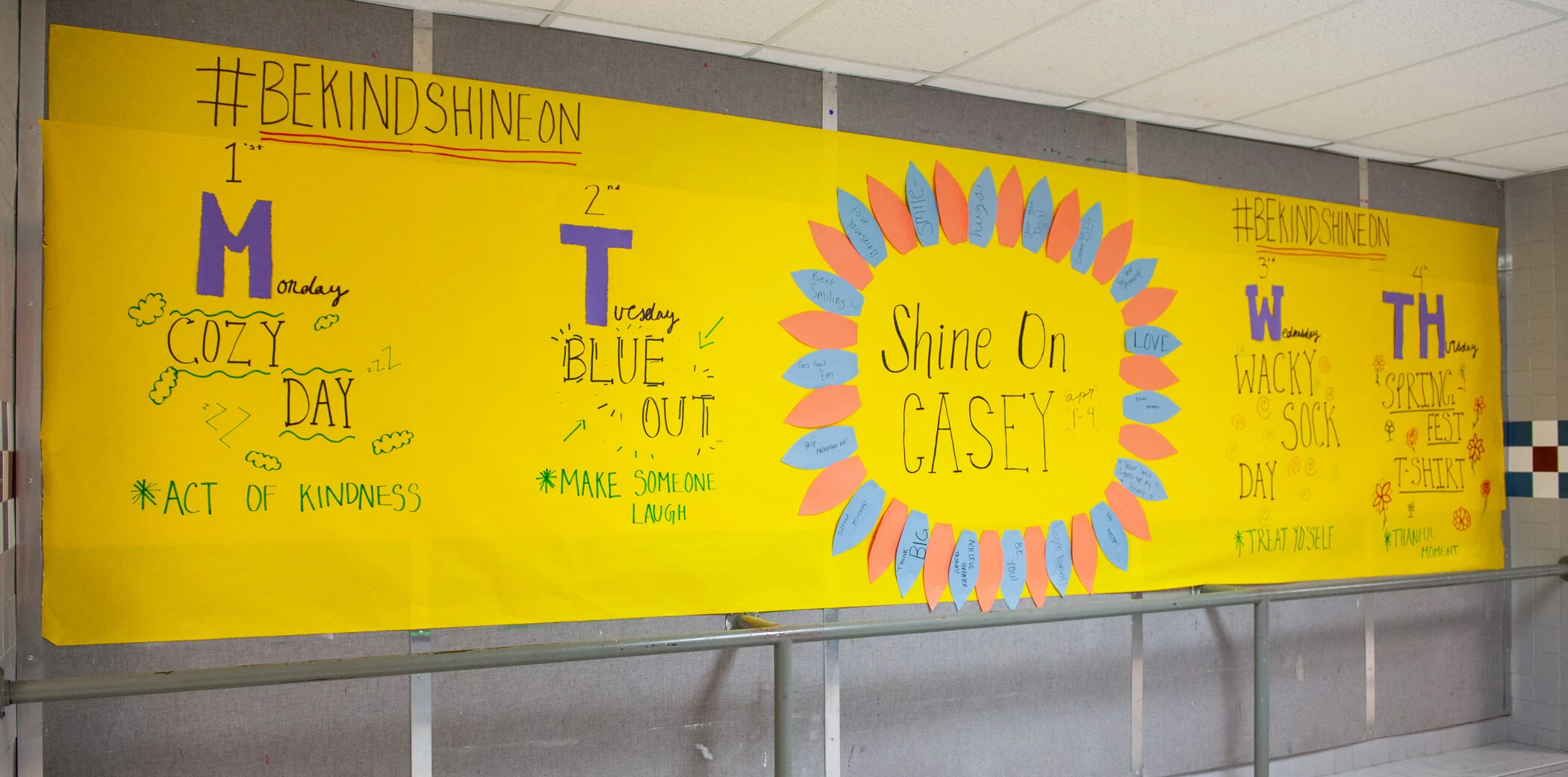 A colorful school bulletin board with the hashtag #BEKINDSHINEON at the top. It highlights daily positivity themes for Monday through Thursday, each with related messages and doodles. Monday promotes 'COZY DAY' with act of kindness, Tuesday features 