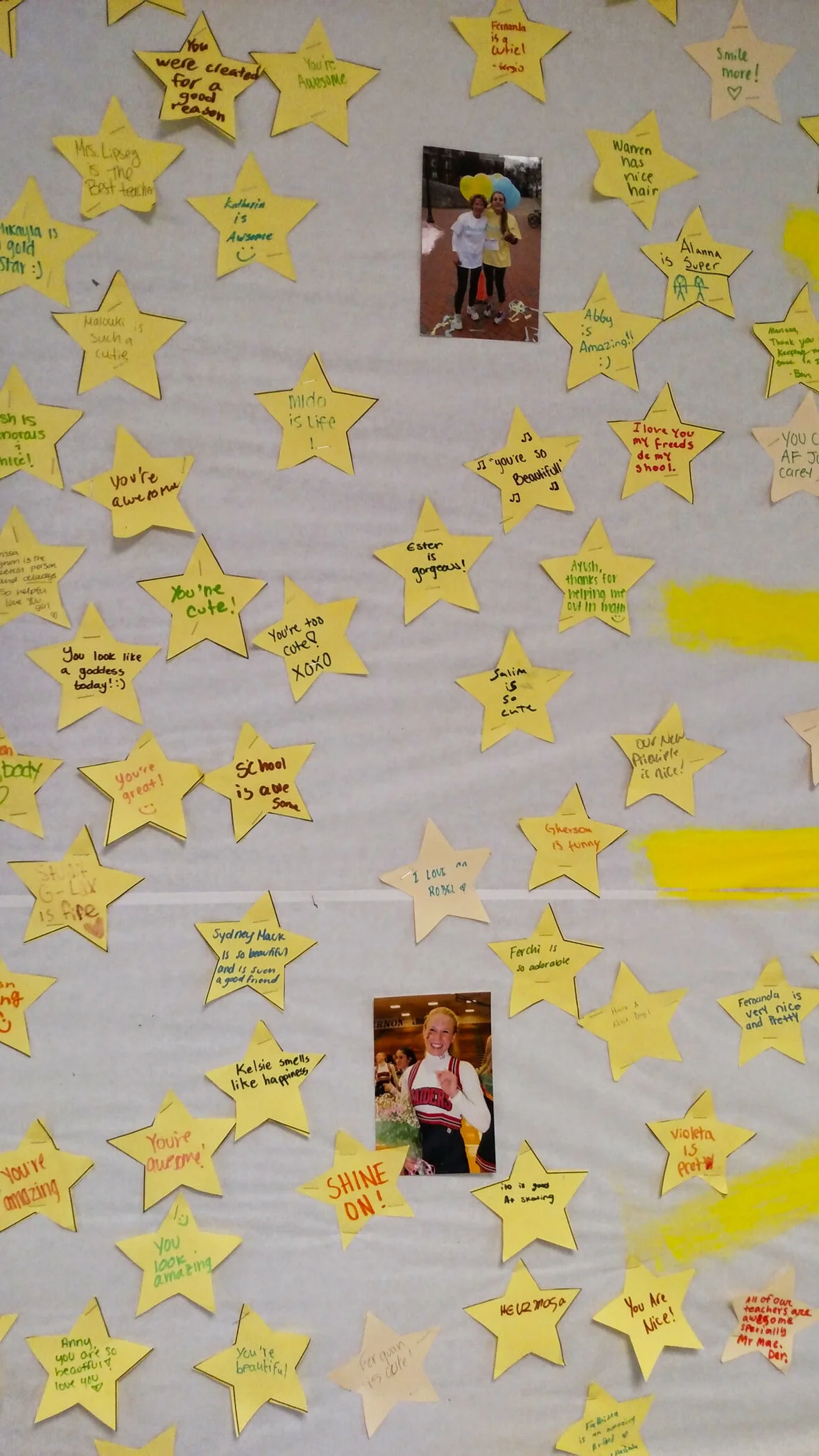 A classroom wall decorated with yellow star-shaped notes containing positive messages and compliments, alongside two photographs of smiling children.