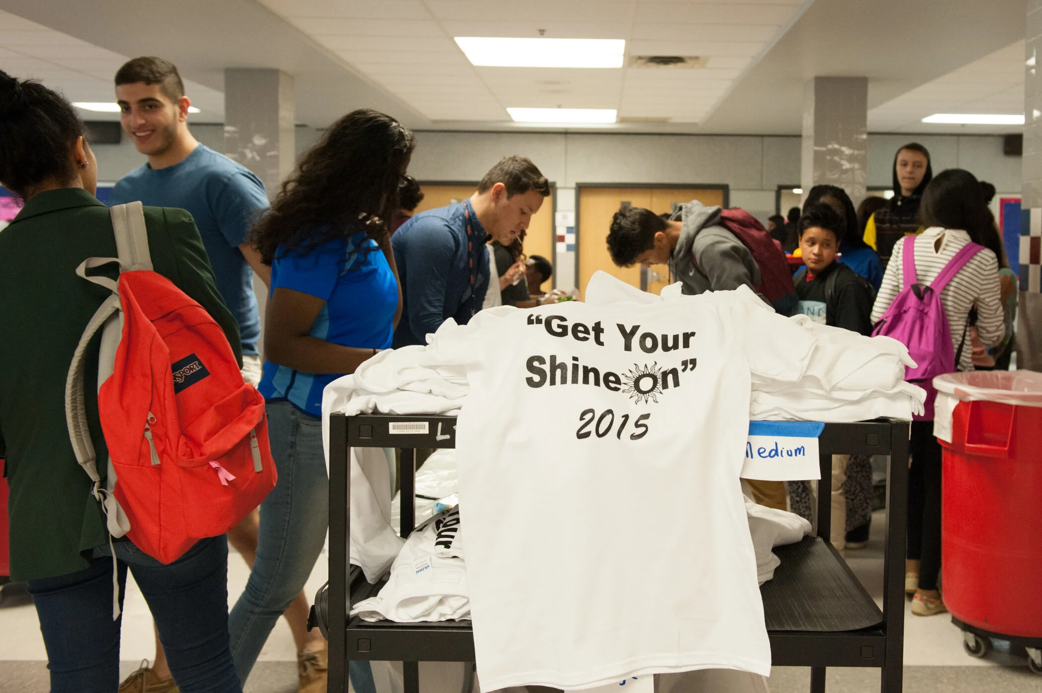 Group of students gathered around a table with white T-shirts that have a slogan 'Get Your Shine On 2015'; some students are looking at the shirts, others are waiting in line.