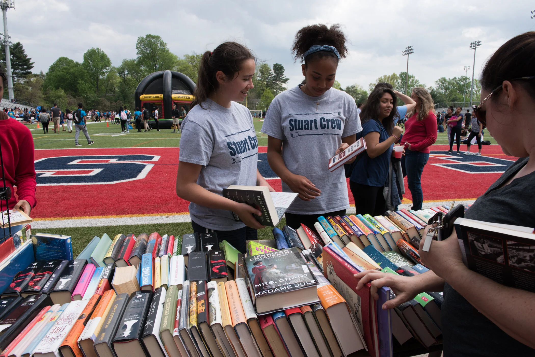 Students browsing books at an outdoor book sale on a school field, with a red and white sports field and other people in the background.