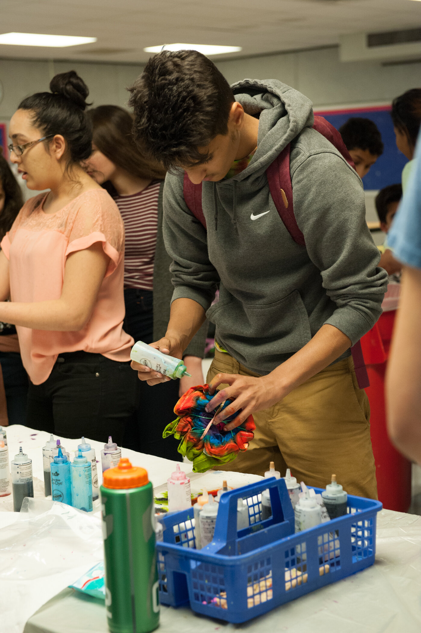 A boy with a gray hoodie and red backpack engaging in tie-dye activity at a table with colorful bottles of dye, surrounded by other people in a classroom or workshop setting.