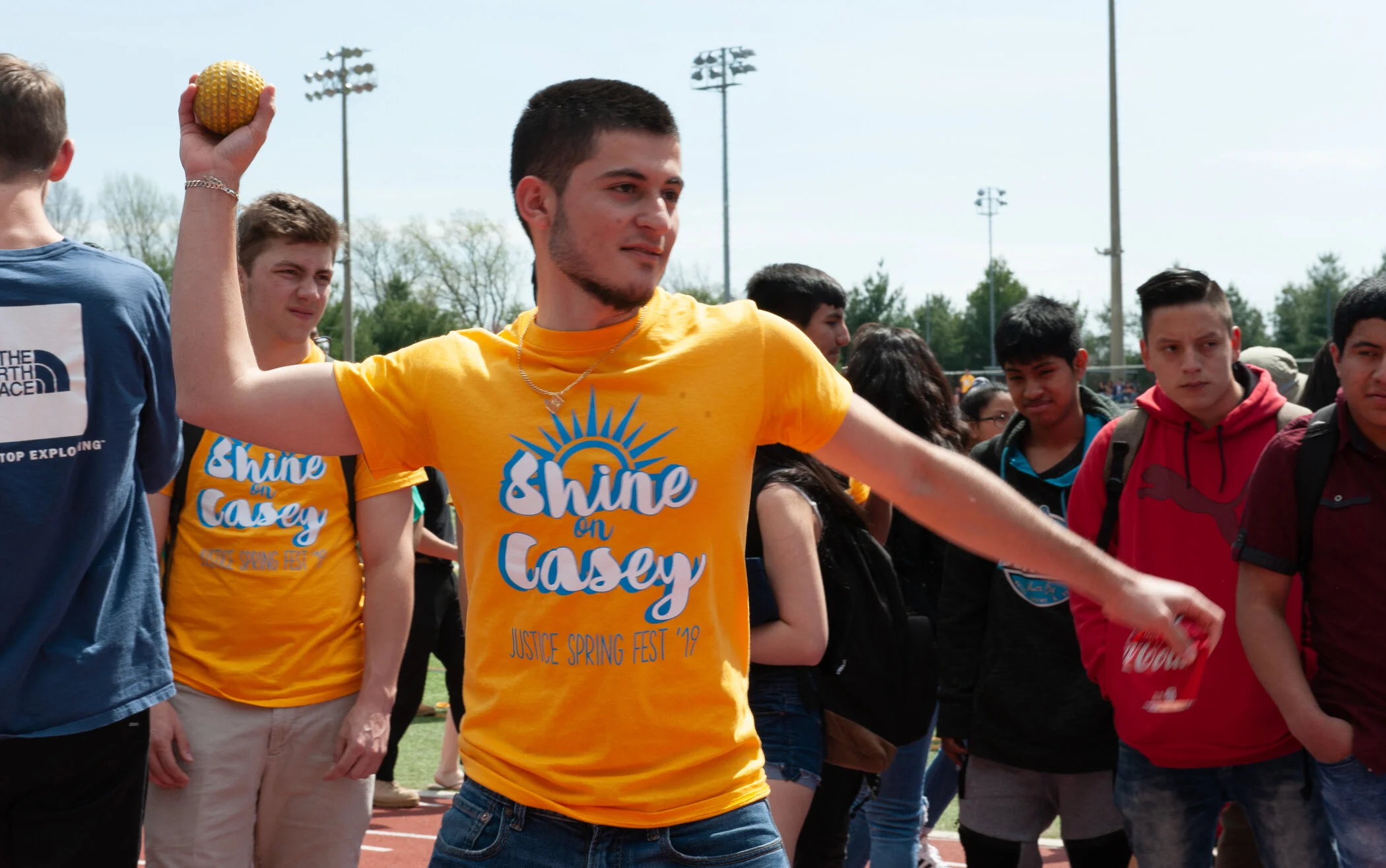 A young man holding a yellow tennis ball on a track field during an event, wearing a yellow T-shirt with the words 'Shine on Casey' and 'Justice Spring Fest 19' written on it, surrounded by other young people.