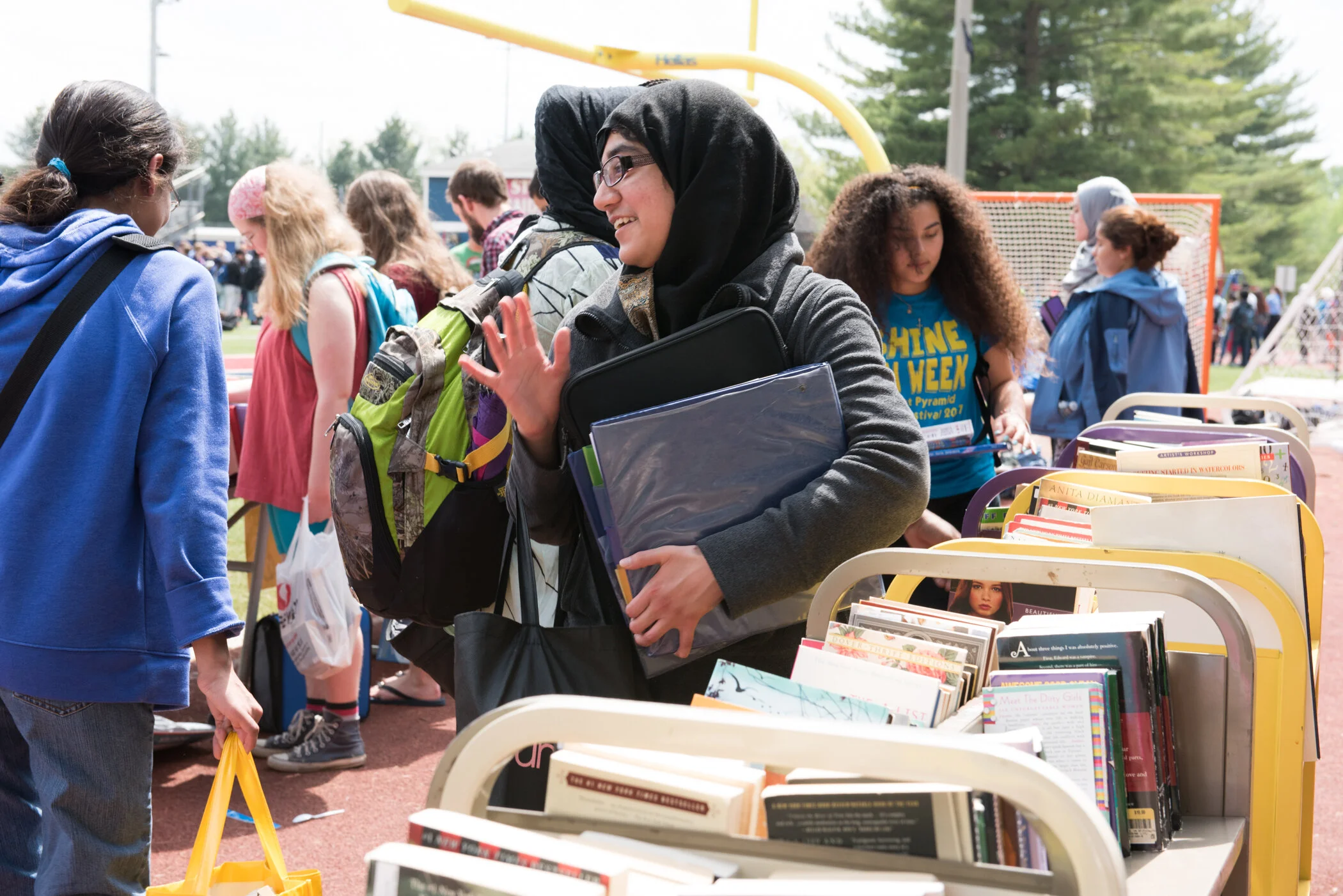 People at a book fair or outdoor market, browsing books on carts, with a woman smiling and waving, wearing glasses, a gray jacket, and a hijab, in a crowd of diverse individuals.