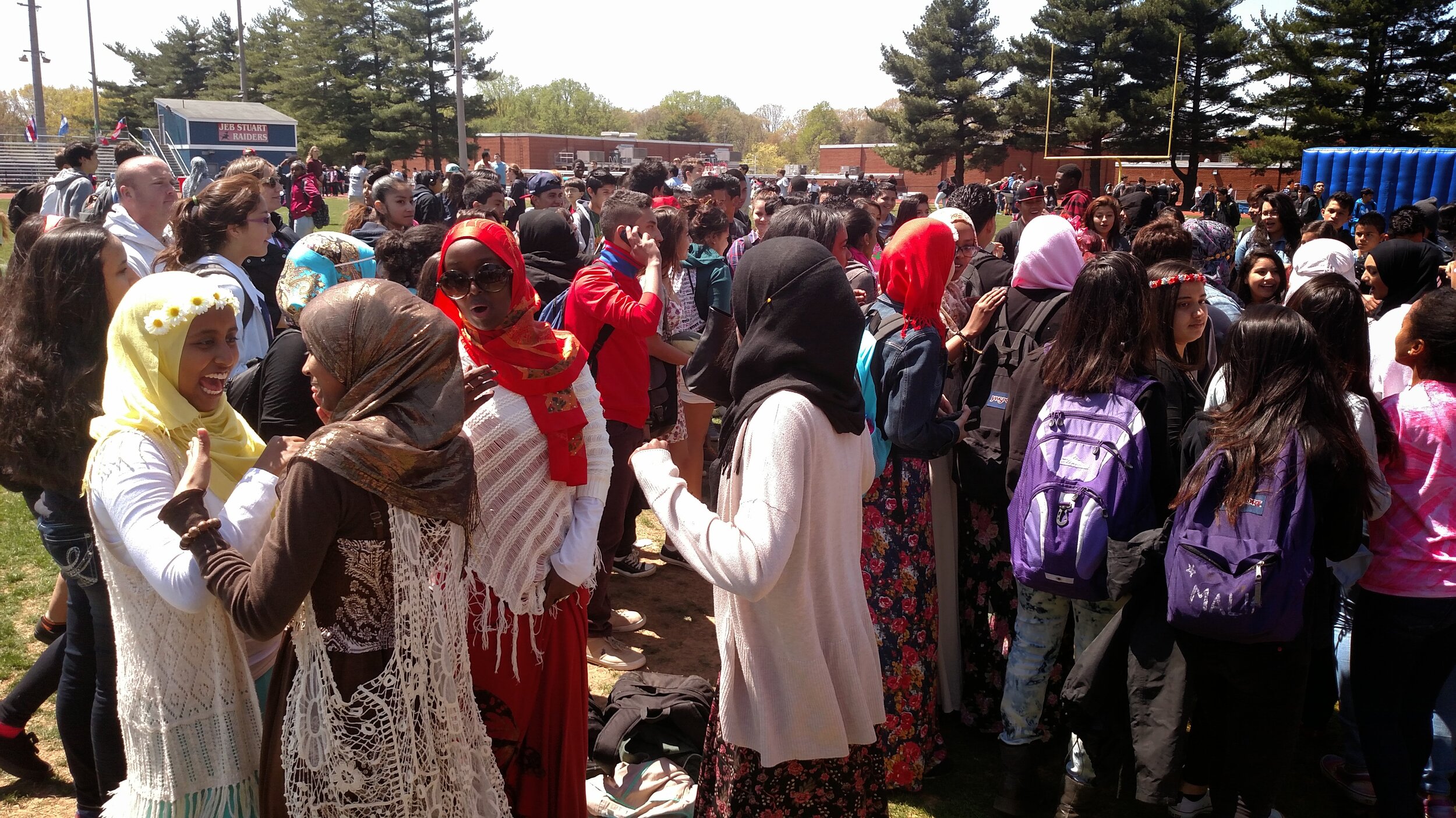 Crowd of diverse students gathered outdoors on a sunny day near a football field, some wearing hijabs and colorful clothing, with a blue inflatable obstacle course and football goalposts in the background.