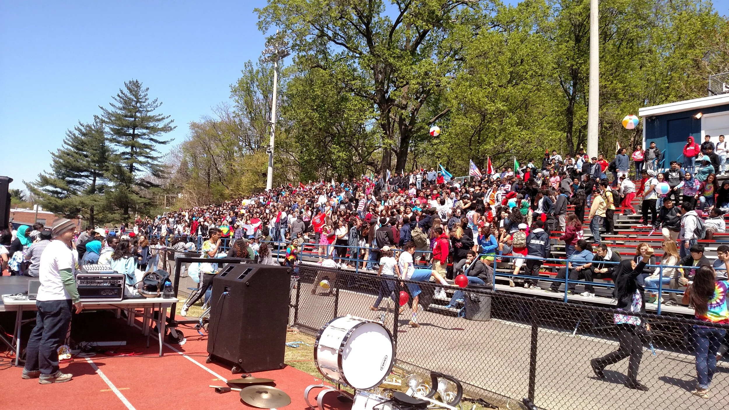 A large outdoor gathering at a school or community event with many young people on bleachers, some waving flags and holding balloons, under a clear blue sky with trees in the background.