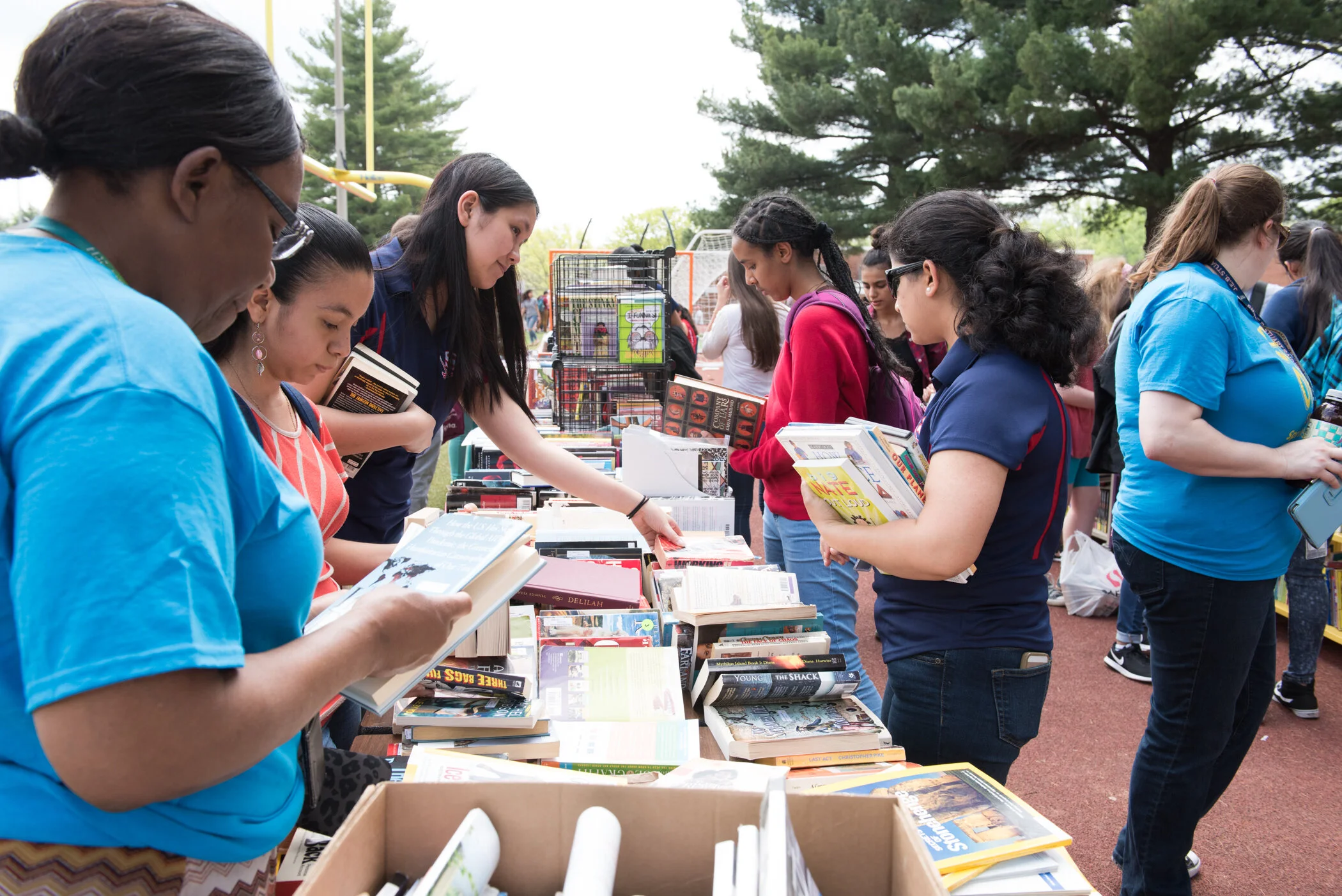 People browsing books at an outdoor book sale or market with tables of books and trees in the background.