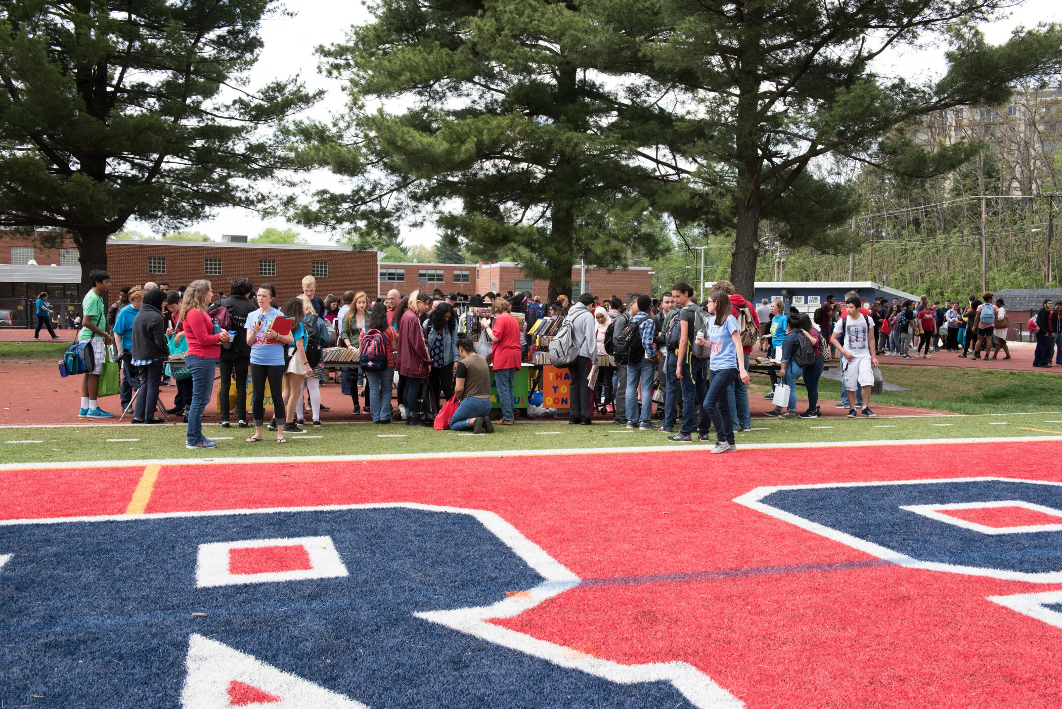 Students gather around tables with books and artwork on a school track field on a cloudy day.
