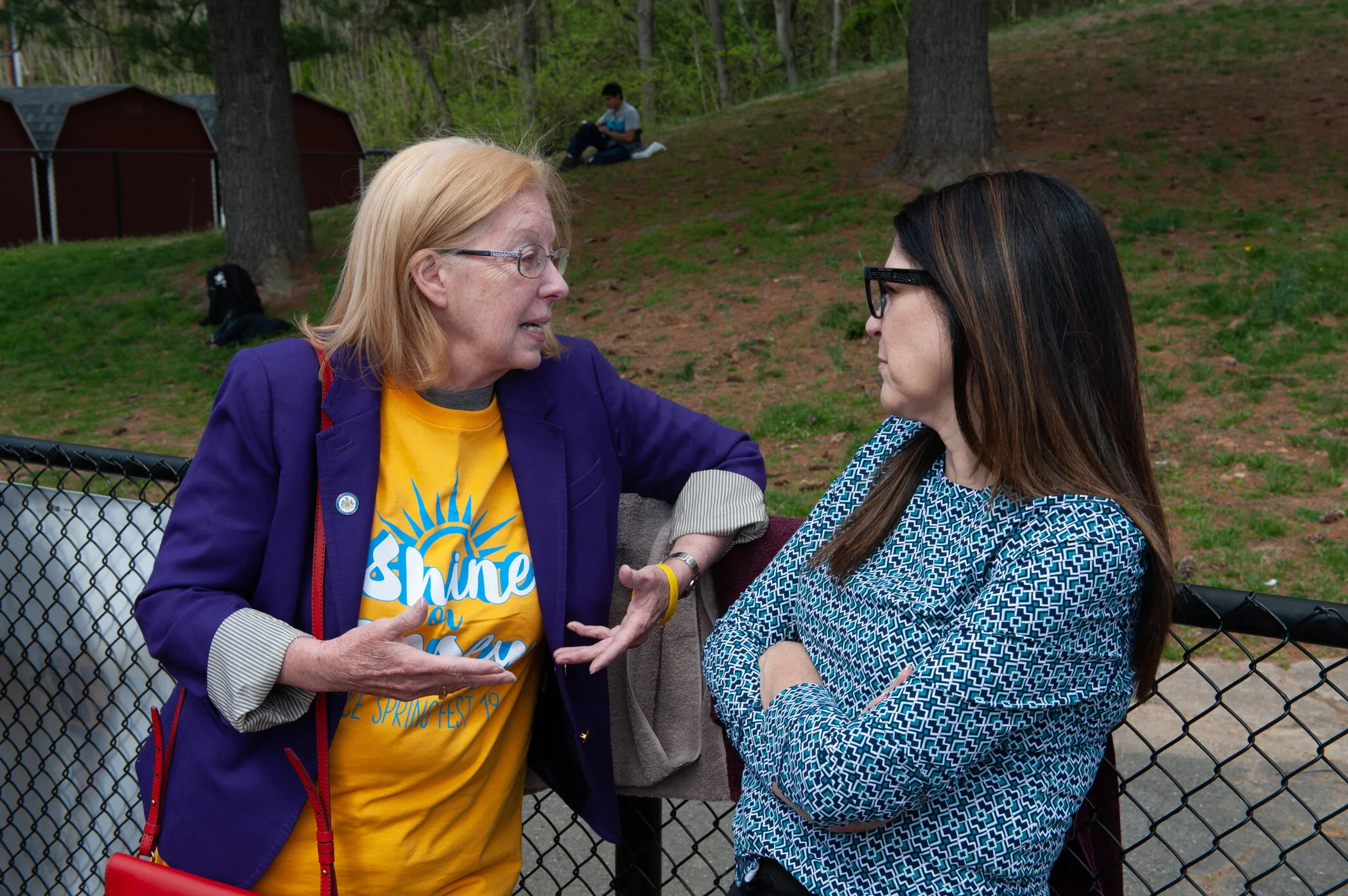 Two women engaged in conversation outdoors, with a young boy sitting on a grassy hill in the background, behind a chain-link fence and trees.