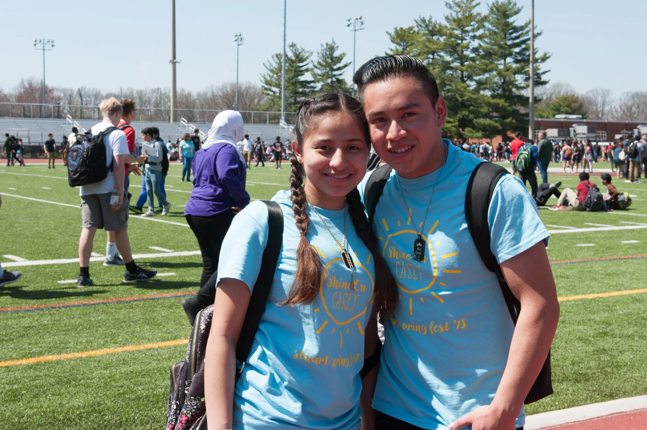 Two students smiling at the camera during a school event on a field, wearing matching light blue t-shirts with yellow text, backpacks, and black dog tags, with many people and students in the background.