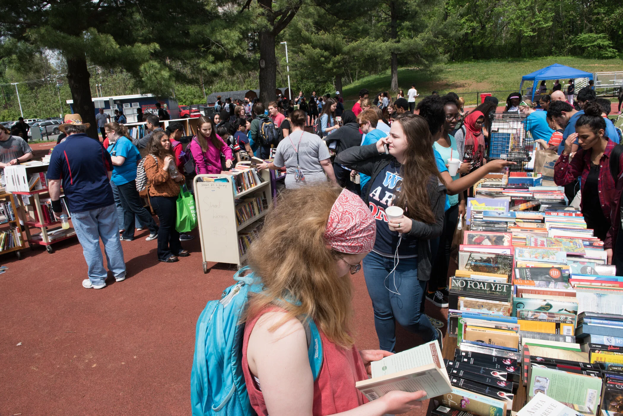 People browsing books at an outdoor book sale, with numerous tables filled with books, and a crowd of people of various ages and backgrounds under trees on a sunny day.