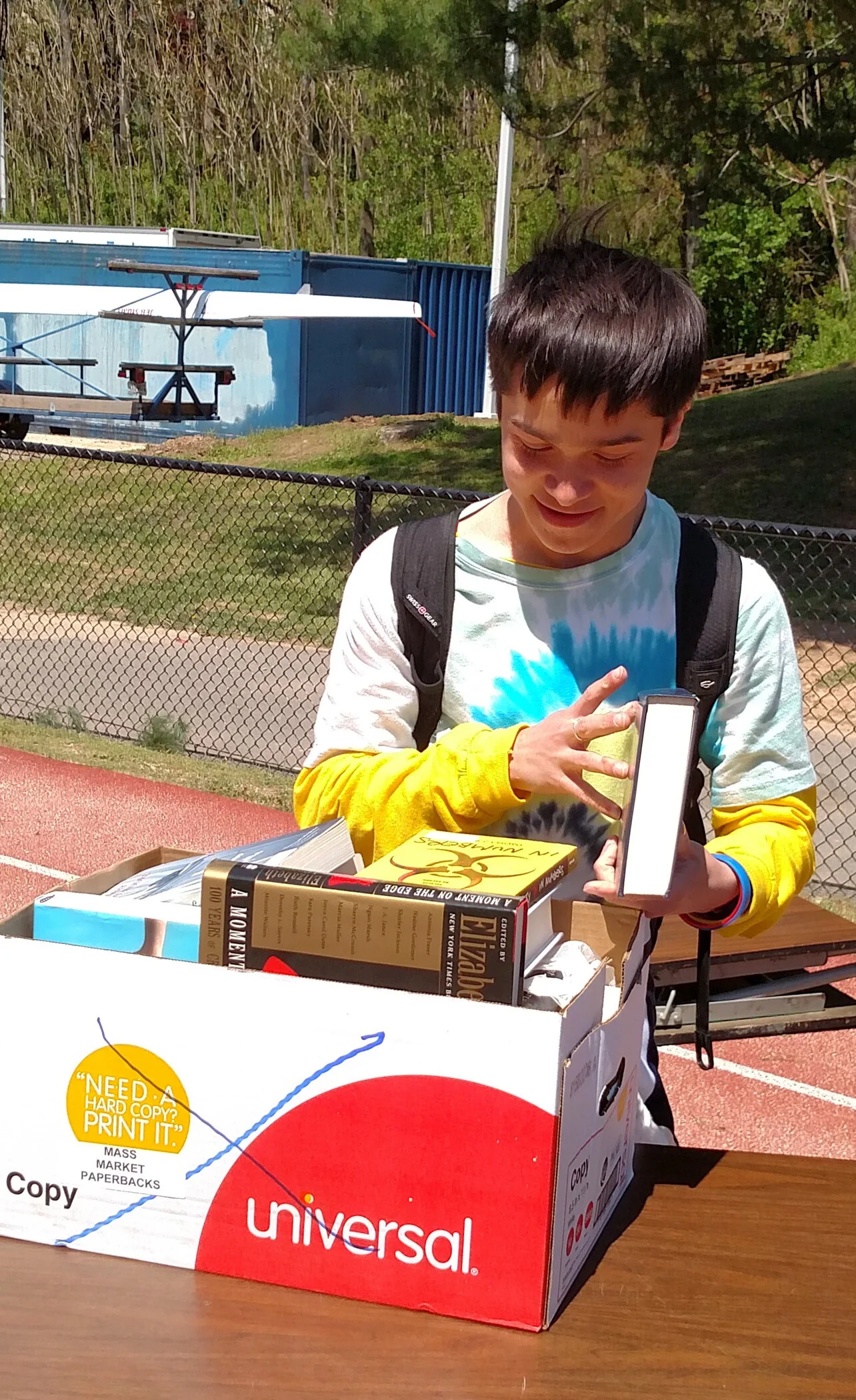 A young boy with a backpack looking at a book inside a box labeled 'Universal' outdoors near a running track, with trees and a fence in the background.