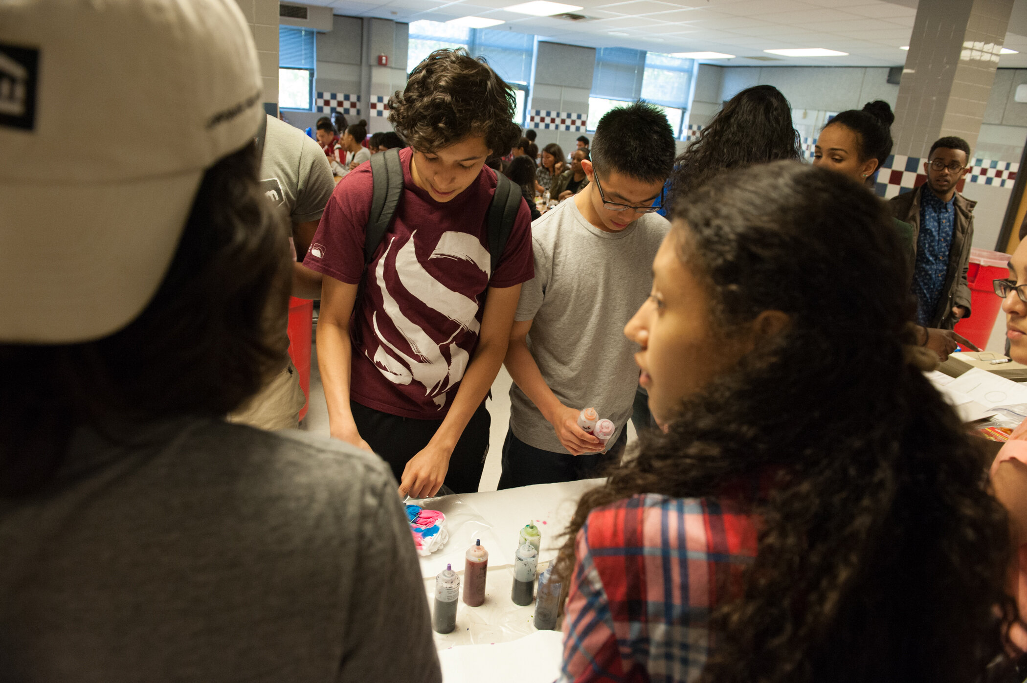 Group of students gathered around a table with colorful paint bottles in a school cafeteria.