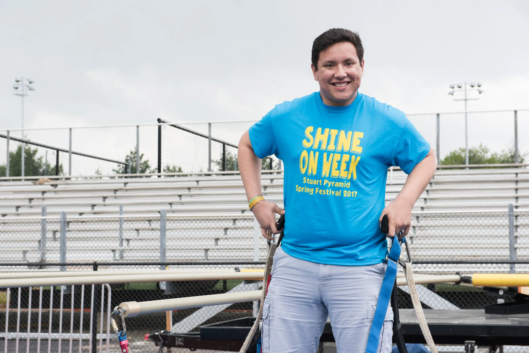 A young man smiling and standing with arms on his hips in an outdoor sports stadium, wearing a blue T-shirt with yellow text that reads 'SHINE ON WEEK, Stuart Pyramid, Spring Festival 2017.'