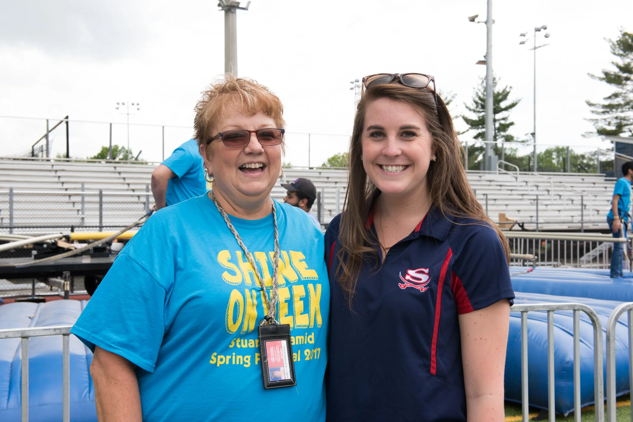 Two women smiling at an outdoor event, surrounded by metal barricades and bleachers, with inflatable obstacles in the background.