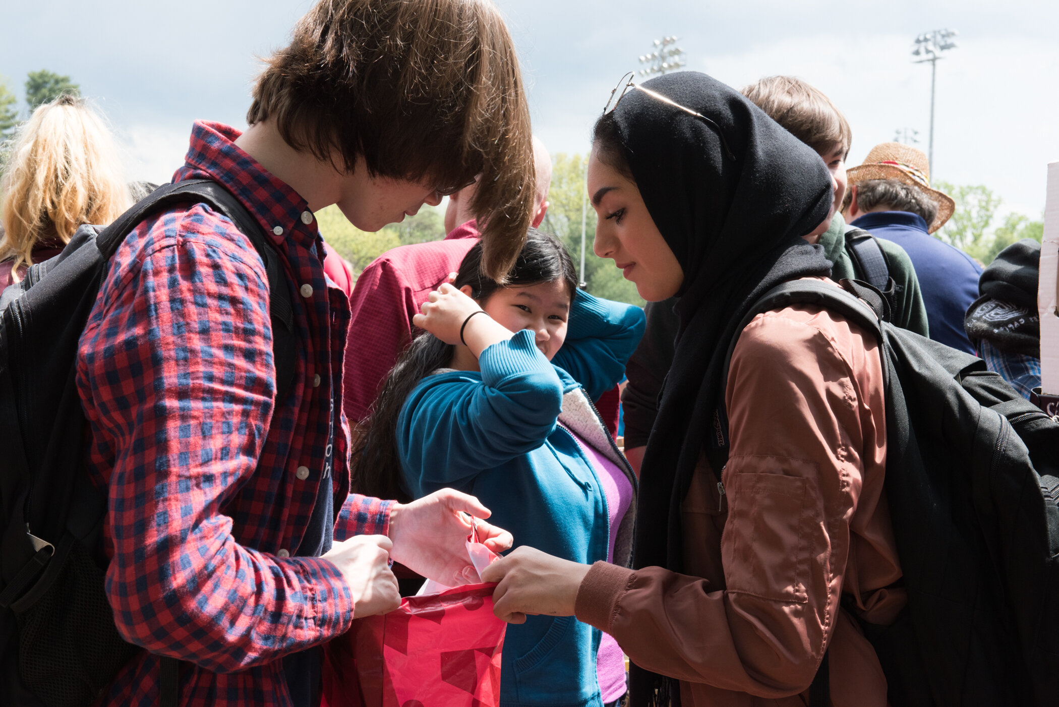 Two young women are exchanging a small item, surrounded by children and adults outdoors in a park or sports field.