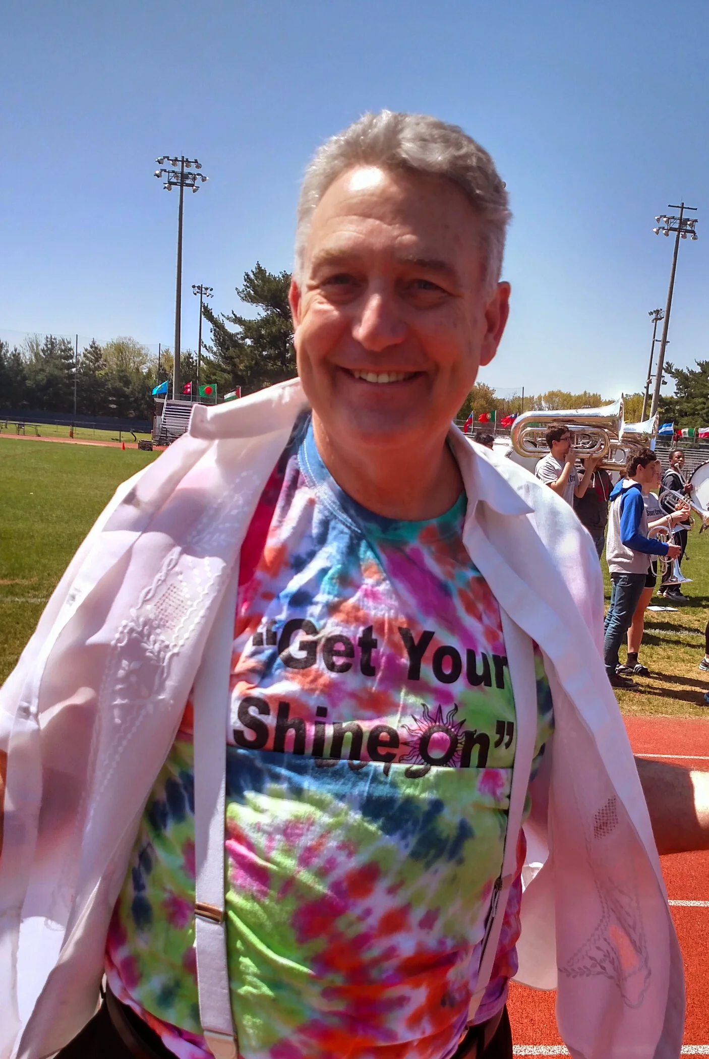 A man smiling at an outdoor event, wearing a tie-dye shirt with the words 'Get Your Shine On' and a white jacket draped over his shoulders. In the background, there are musicians playing brass instruments on a field, with a track and sports field equ