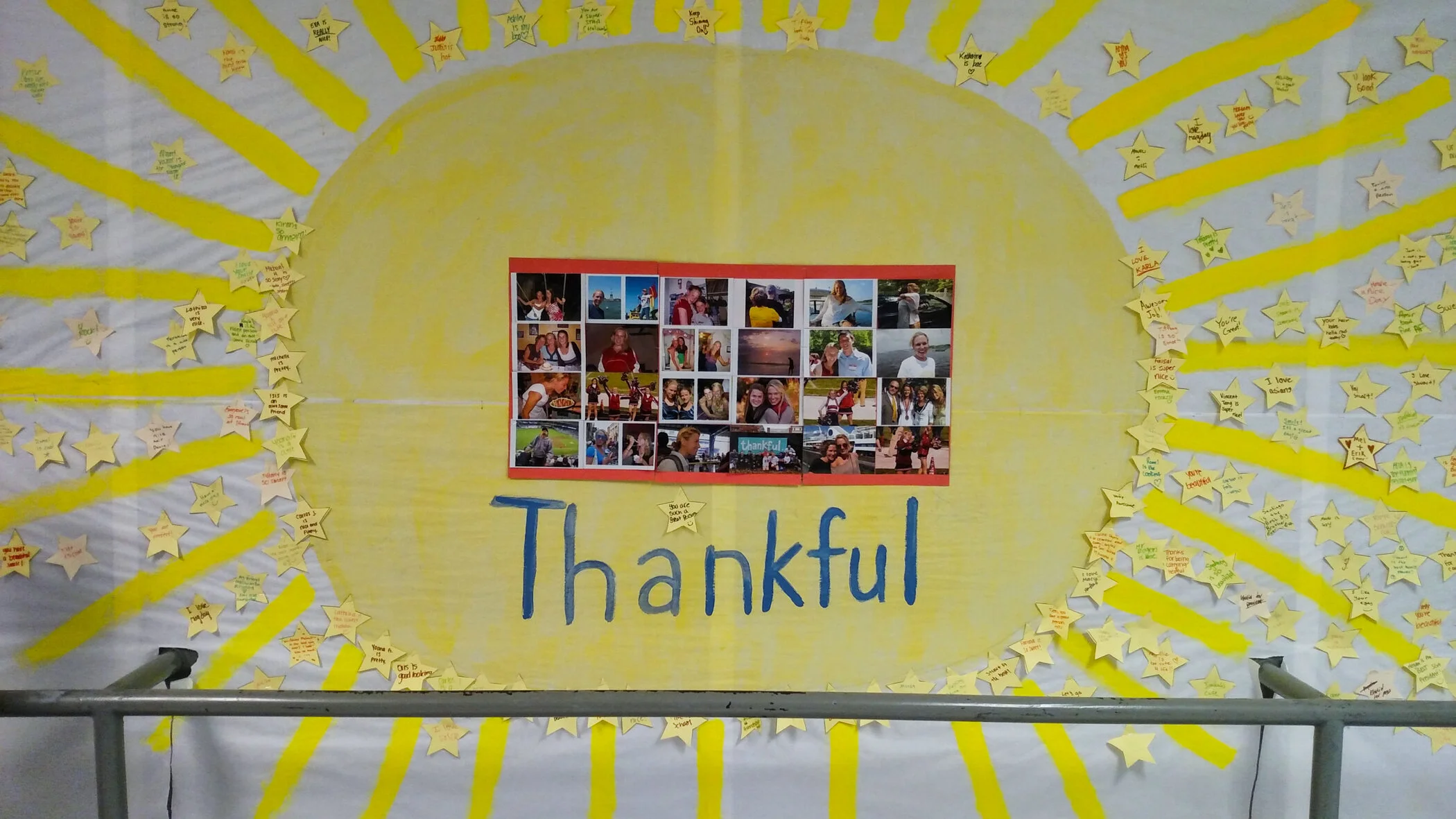 A large display with a yellow background and yellow rays emanating outward, decorated with numerous yellow star-shaped notes containing handwritten messages. In the center, there is a collage of photographs showing various people and activities. Belo