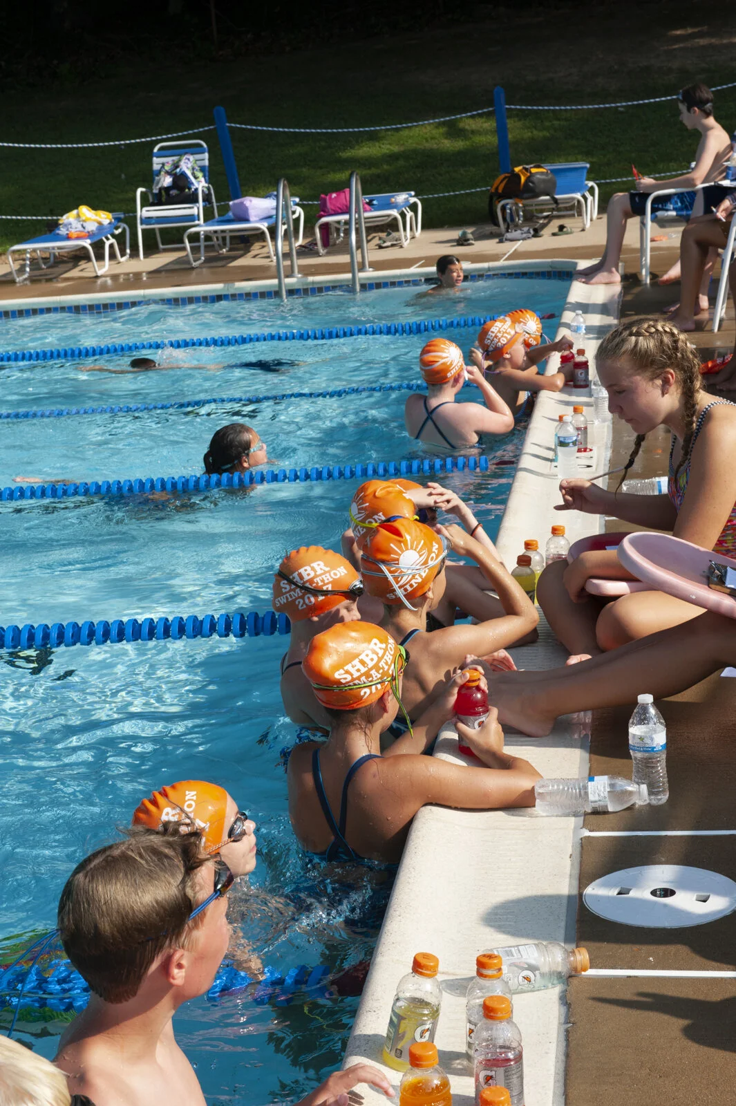 Children and teenagers at a swimming pool during a swim meet or practice, some wearing orange swim caps, others in swimsuits, sitting on the pool deck, and some swimming in the lanes.