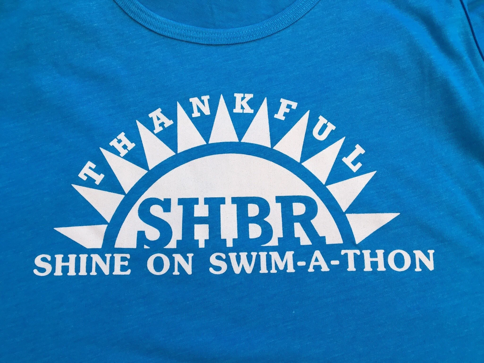 A blue T-shirt with a design that features a white sun with rays and text that reads, 'The Thankful SHBR Shine on Swim-A-Thon'.