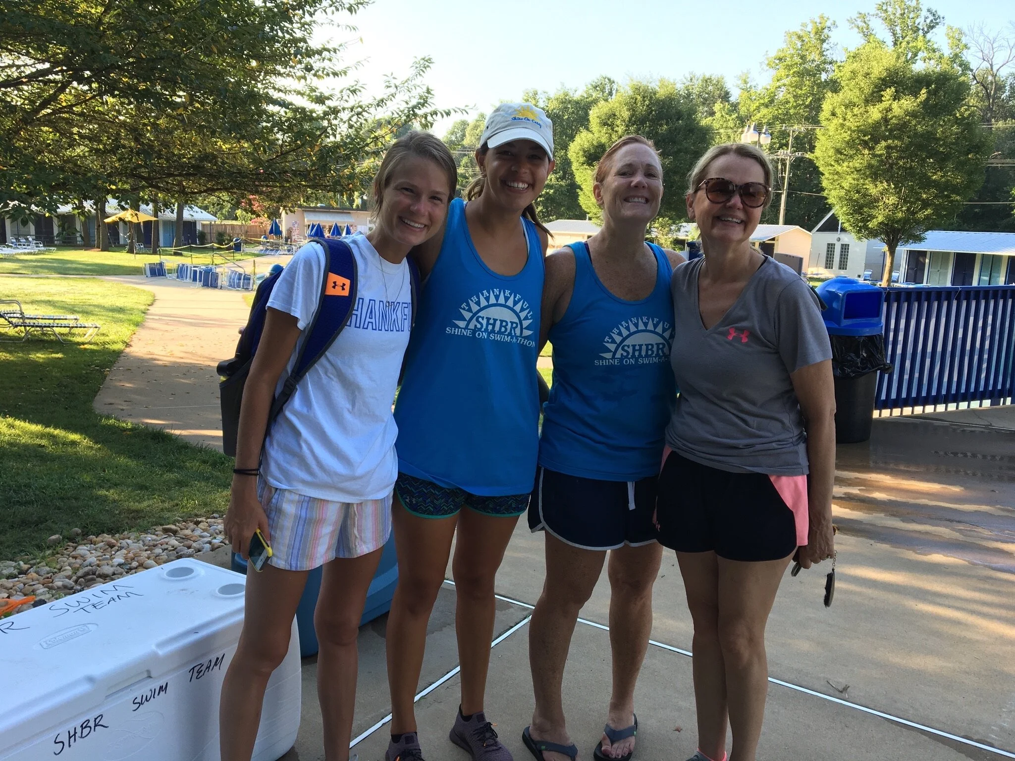 Four women standing outdoors near a swimming pool, smiling for the photo. One woman is wearing a white shirt and striped shorts with a backpack, the second and third women are in blue shirts with a swim team logo, and the fourth woman is in a gray sh