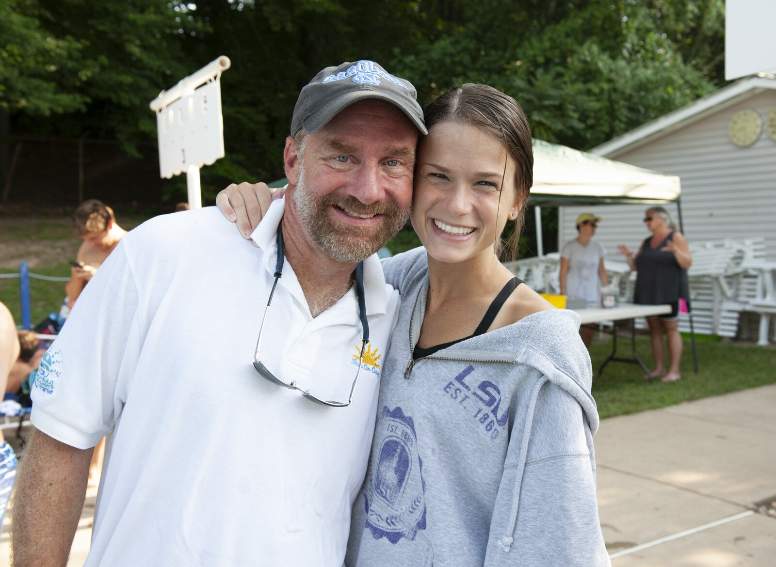 A smiling man and woman standing outdoors at a gathering, with trees and a building in the background, and other people in the distance.